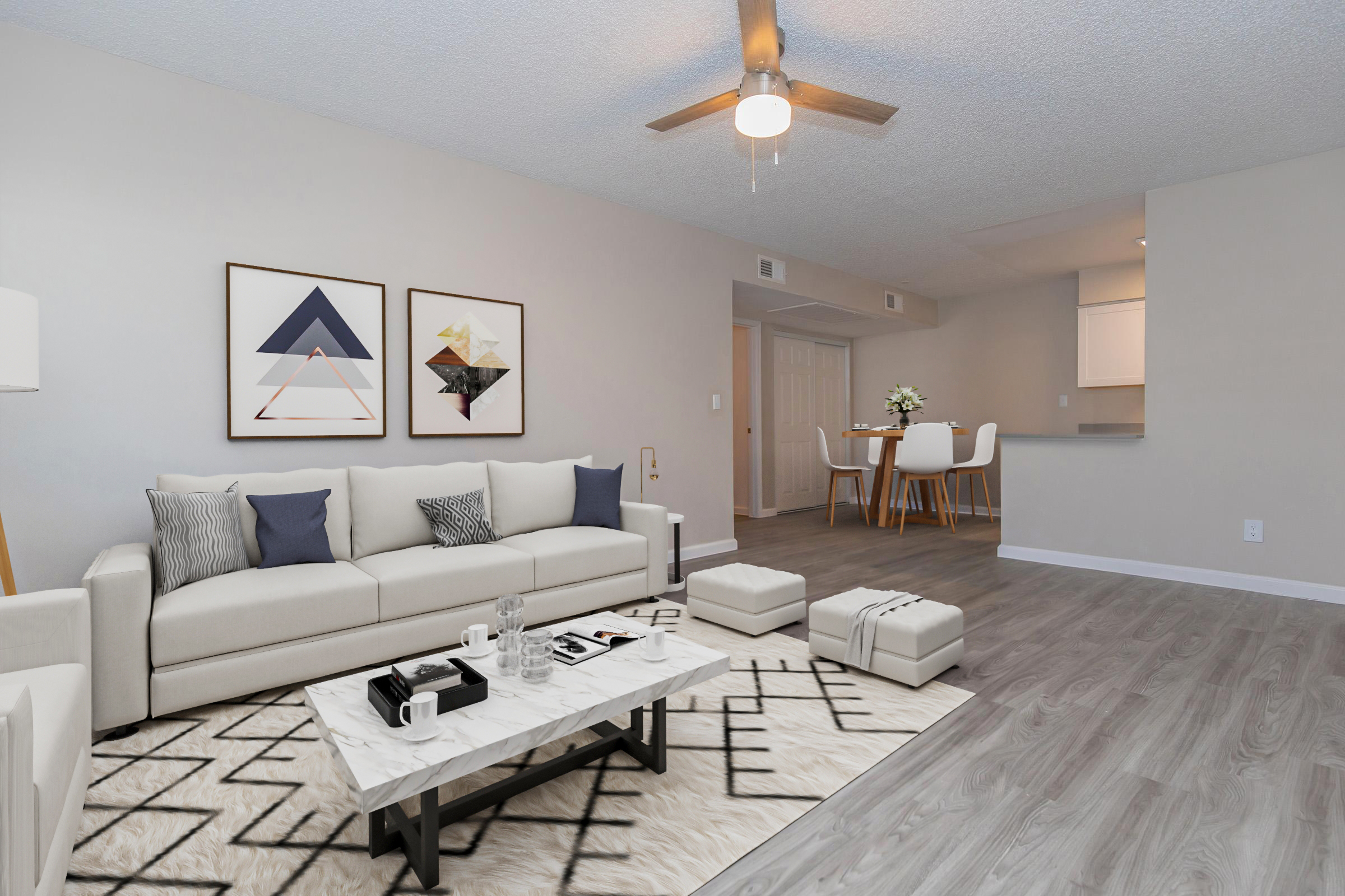 A modern living room featuring a light-colored sofa with decorative pillows, a geometric coffee table, and a patterned area rug. Wall art with abstract designs hangs above the sofa. A dining area with a light wooden table and four chairs is visible in the background, along with a ceiling fan.