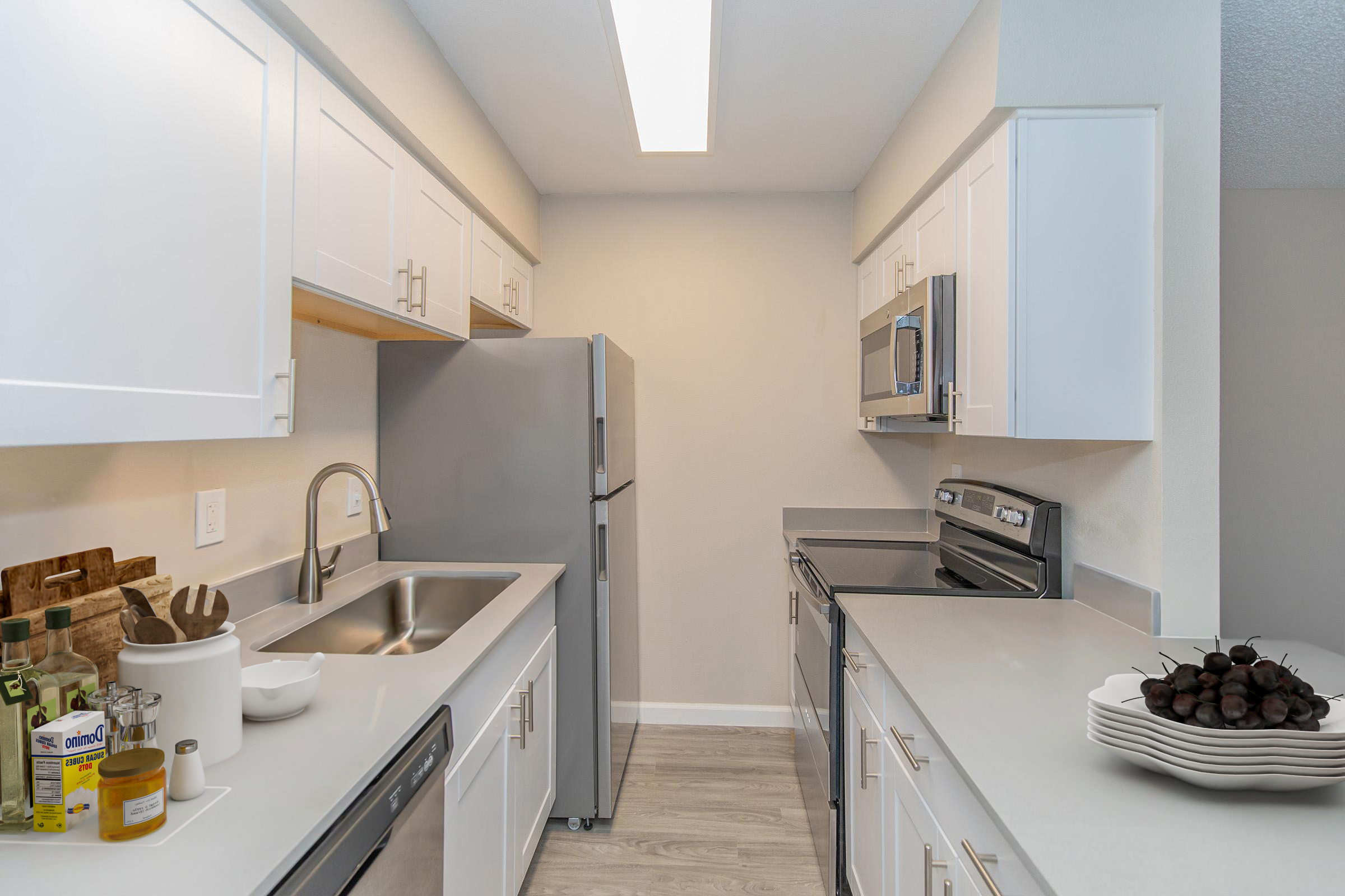 Modern kitchen with light-colored cabinetry and stainless steel appliances. Features a double sink, electric range, and refrigerator. The countertops are sleek and clean, with a decorative white bowl of fruits on the right. The overall aesthetic is bright and inviting, with a minimalist design.
