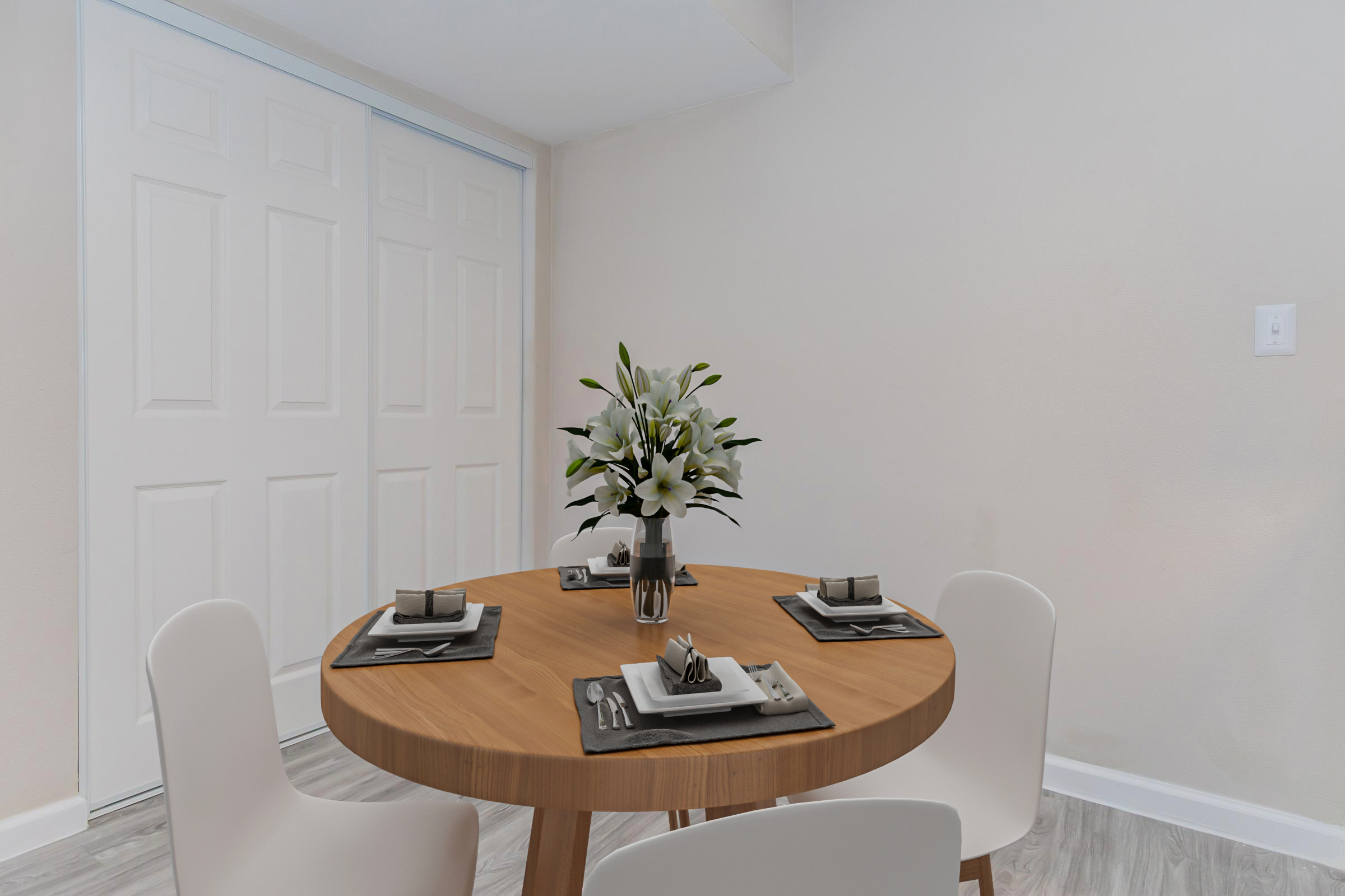 A modern dining area featuring a round wooden table set for four with black and white place settings. In the center, a vase with white flowers adds a touch of elegance. The background shows a pair of closed white double doors and light-colored walls, creating a bright, inviting atmosphere.