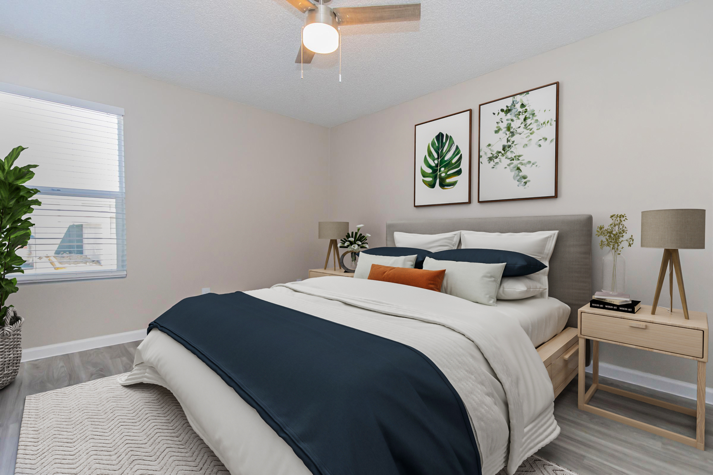 A cozy bedroom featuring a bed with a navy blue and white bedding set, complemented by decorative pillows. Two bedside lamps sit on wooden nightstands, and artwork of plants hangs on the wall. A potted plant adds a touch of greenery, and a soft rug covers the floor beneath. Natural light filters through a window.