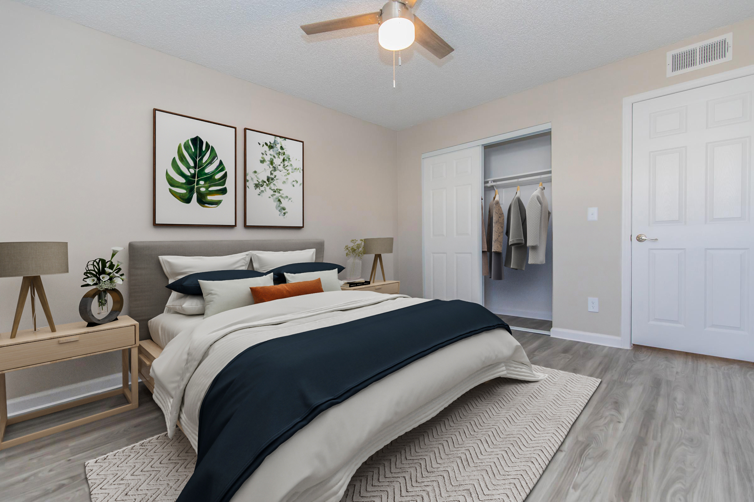 A beautifully arranged bedroom featuring a neatly made bed with navy and beige bedding, two decorative pillows, and framed botanical artwork on the wall. A stylish nightstand with a lamp sits beside the bed, and a closet with hanging clothes is visible in the background. Natural light illuminates the space.