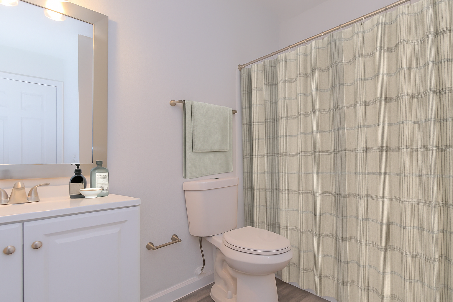 A clean, well-lit bathroom featuring a white toilet, a light-colored shower curtain with a checkered pattern, a wall-mounted towel holder with a folded towel, and a vanity with a mirror and sink. Hand soap and a small dish are on the counter. The walls are painted a soft, neutral color.