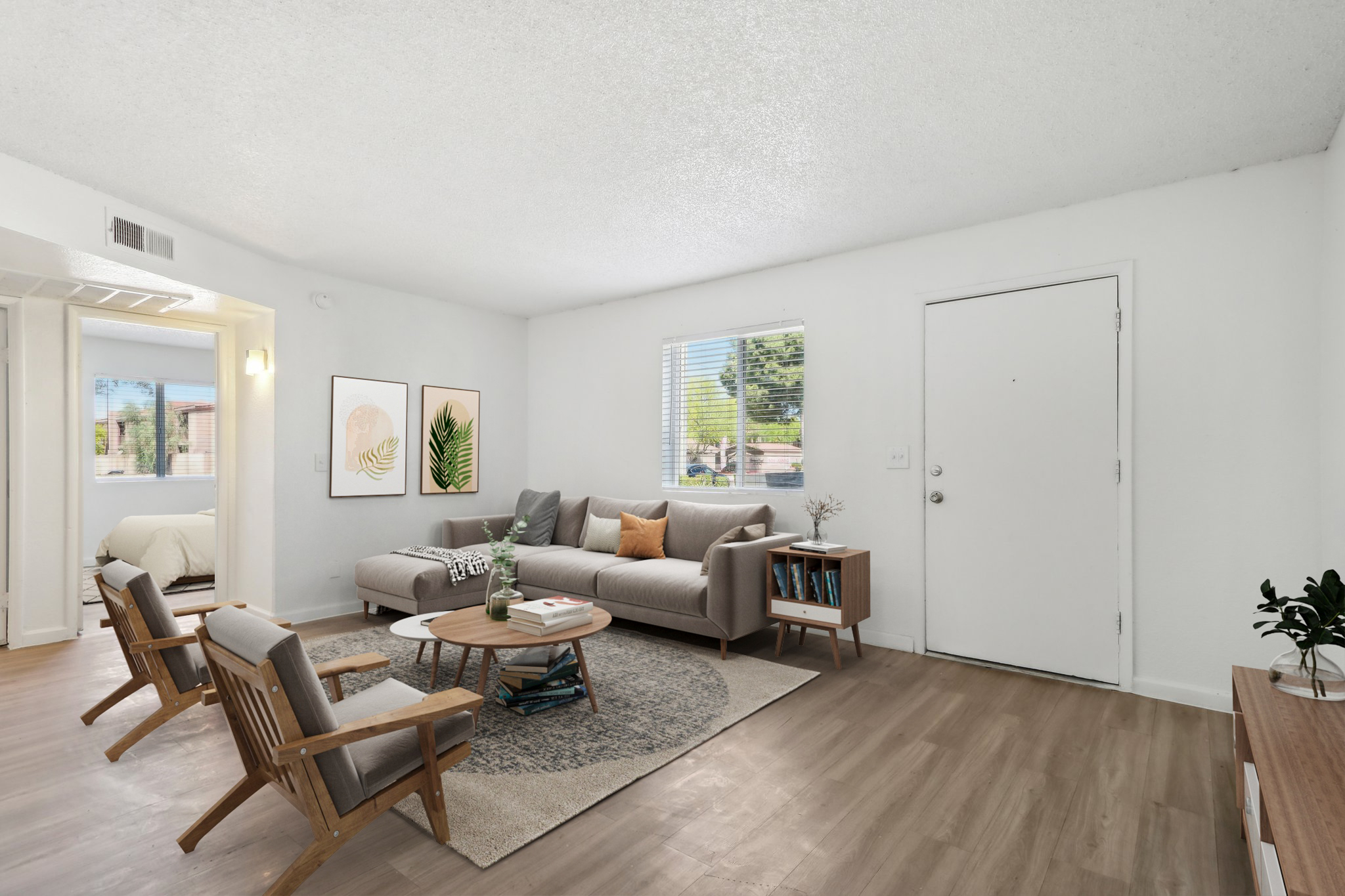 A bright and airy living room featuring a gray sofa with decorative pillows, two wooden chairs, a round coffee table, and a stylish rug. The walls are painted white, and there are framed artworks on display. A large window provides natural light, and a door is visible on the right side of the image.