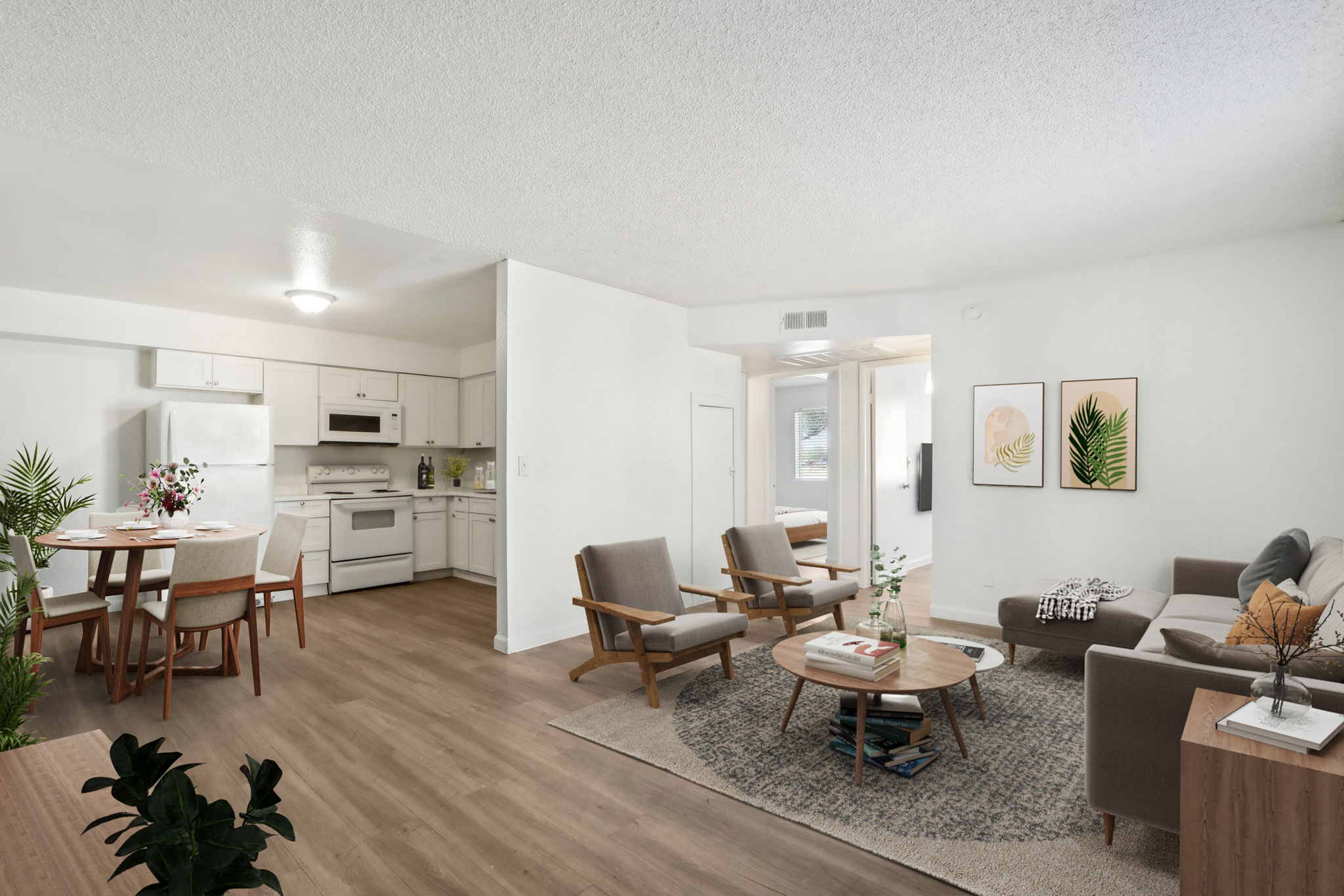 Bright, modern open-concept living space featuring a cozy seating area with a gray couch and wooden chairs, a circular coffee table, and potted plants. In the background, a dining area with a round table and white kitchen with appliances is visible. Light-colored walls and flooring enhance the airy feel.