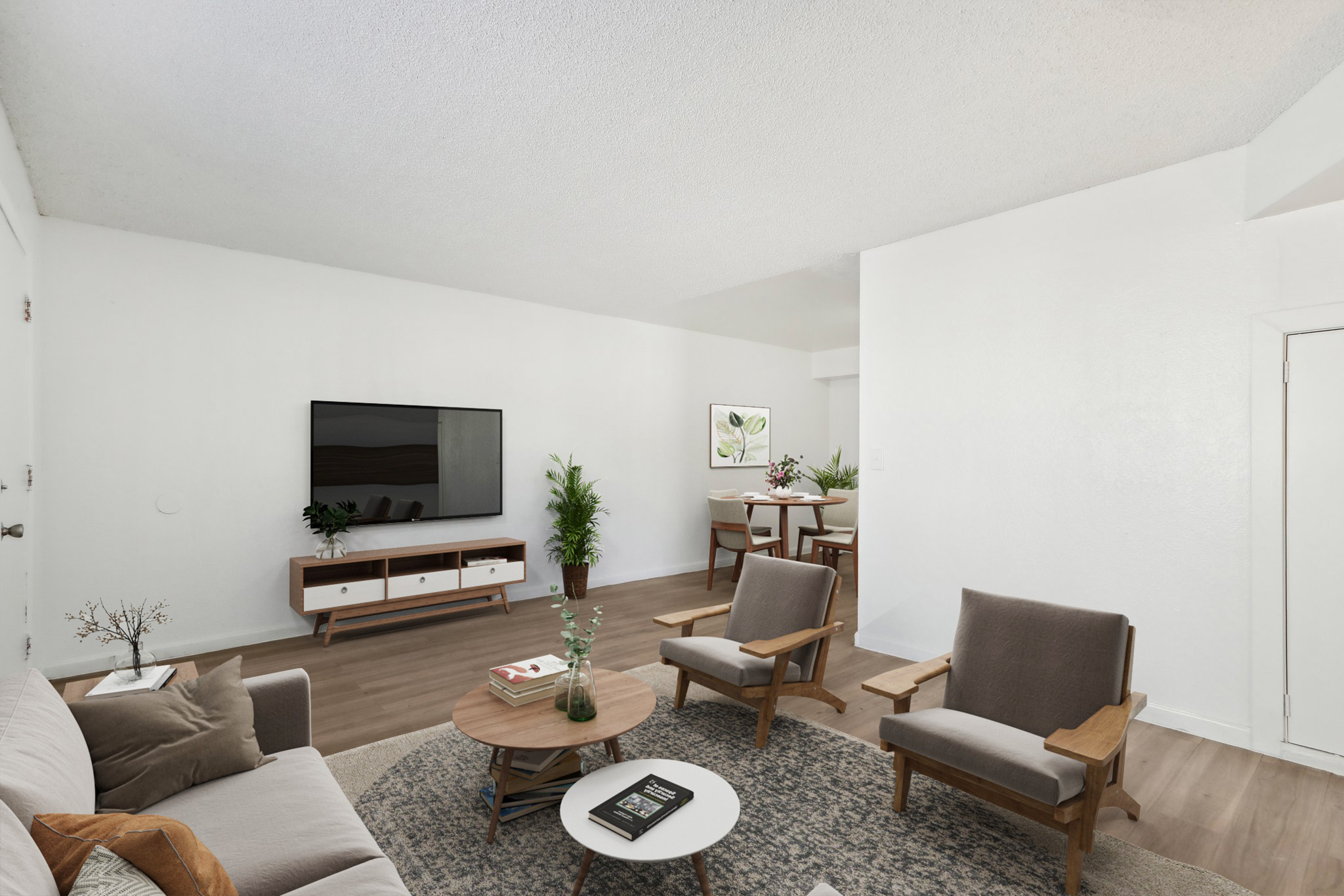 A modern living room featuring a light-colored sofa, two wooden armchairs, and a circular coffee table. A large TV is mounted on the wall above a contemporary media console. There are houseplants in the corners and a dining area visible in the background, all set in a bright, minimalist space.