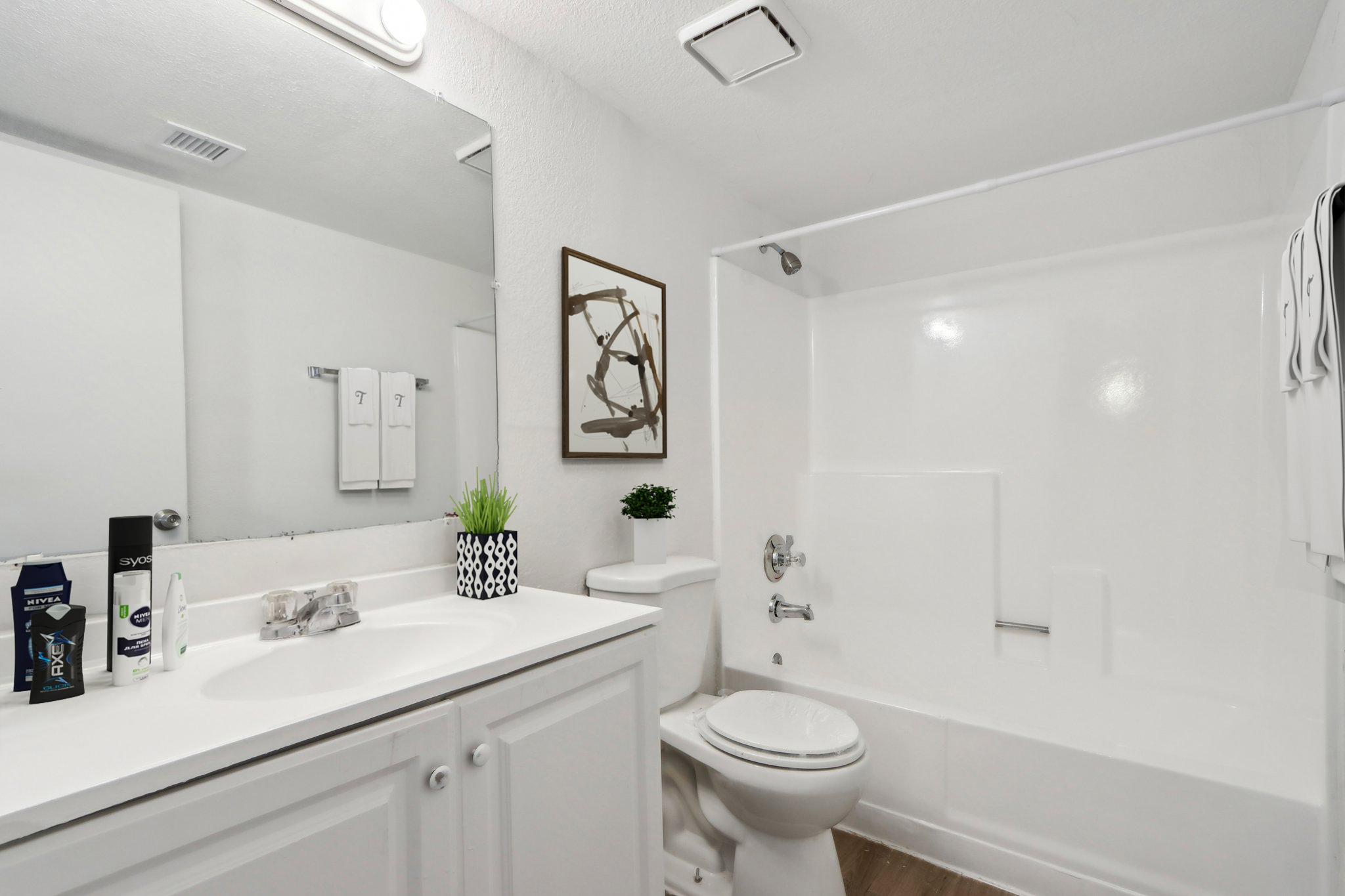 A clean and modern bathroom featuring a white tub and shower, a toilet, and a vanity with a sink. There are two white towels neatly hanging, a small potted plant, and toiletries on the countertop. A decorative framed piece is mounted on the wall above the sink. The overall decor is bright and minimalistic.