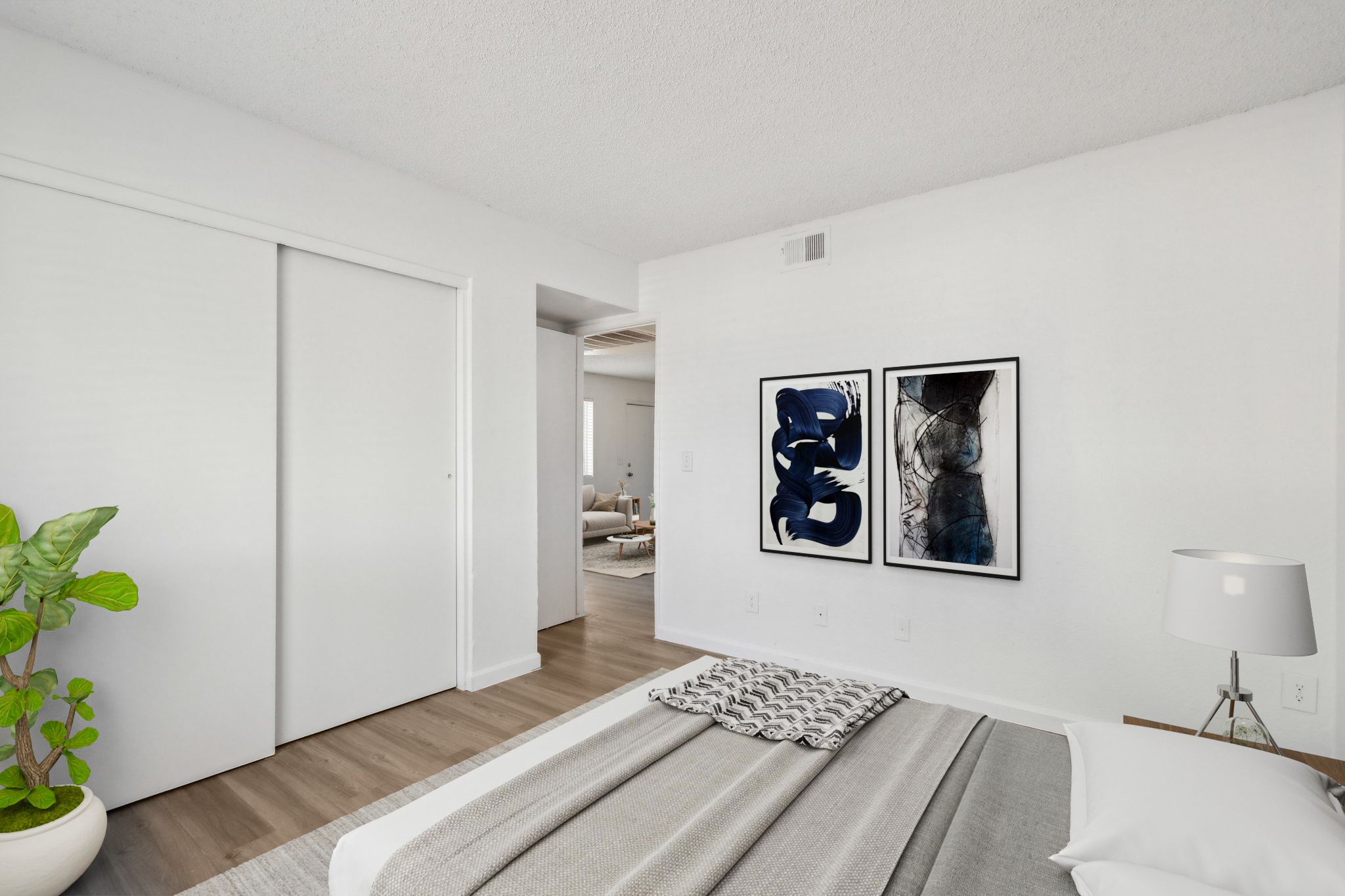 A modern bedroom featuring a neatly made bed with a gray and white blanket, a bedside lamp, and a decorative plant. On the wall, two framed artworks in shades of blue and black add a stylish touch. The room has light-colored walls and wooden flooring, creating a bright and inviting atmosphere.