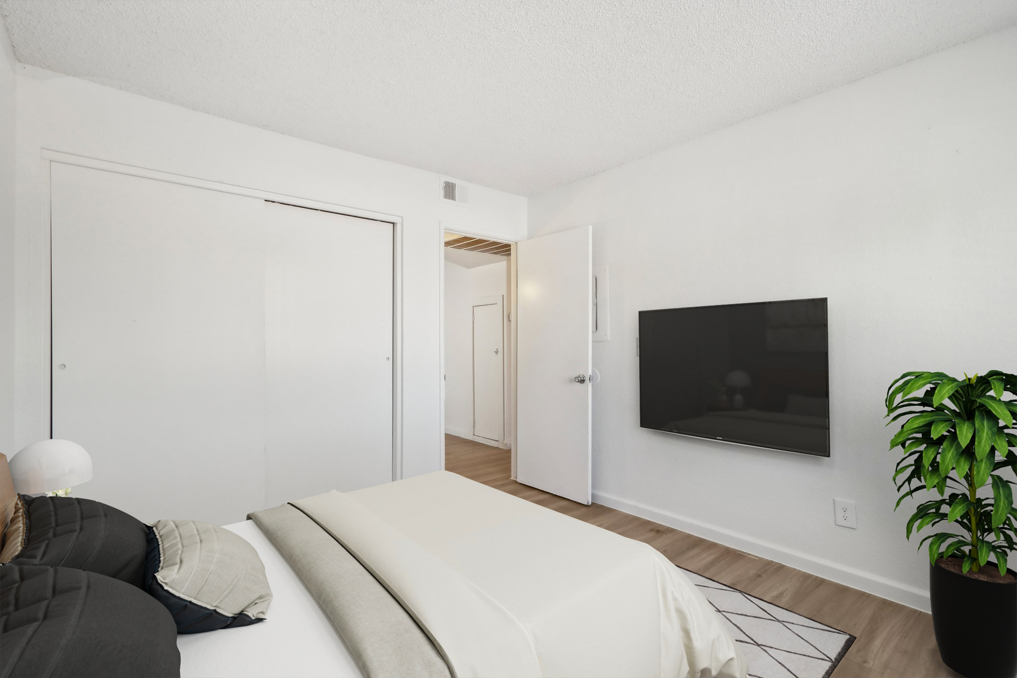 A stylish bedroom featuring a neatly made bed with decorative pillows, a large wall-mounted flat-screen TV, and a potted plant. The room has light-colored walls, wooden flooring, and a closet with sliding doors, creating a modern and inviting atmosphere.