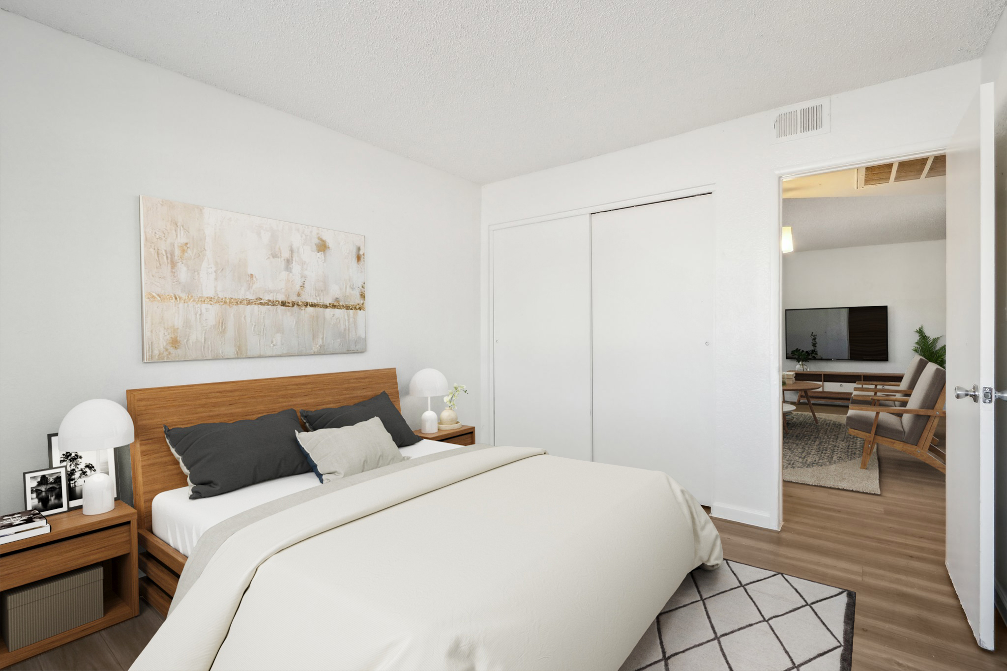 A well-lit bedroom featuring a bed with a neutral-colored blanket and decorative pillows. There are two nightstands with lamps, a piece of abstract art on the wall, and a closet with sliding doors. In the background, a doorway leads to another room with a modern sitting area.