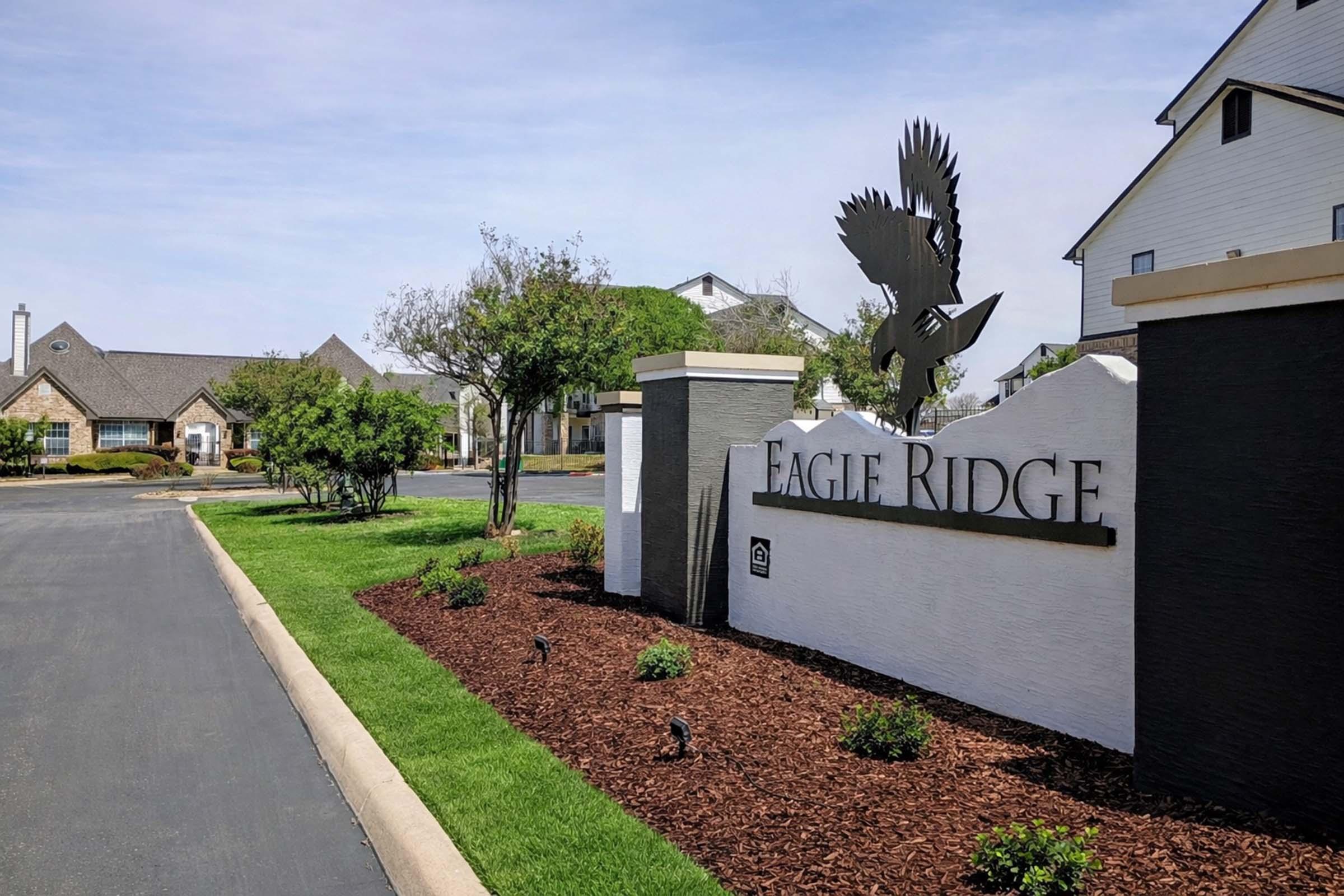 A sign displaying "Eagle Ridge" is prominently placed at the entrance of a residential community. The sign features an eagle silhouette above the text. Surrounding the sign are landscaped areas with green grass, small plants, and mulch, indicative of a well-maintained neighborhood, with buildings visible in the background.