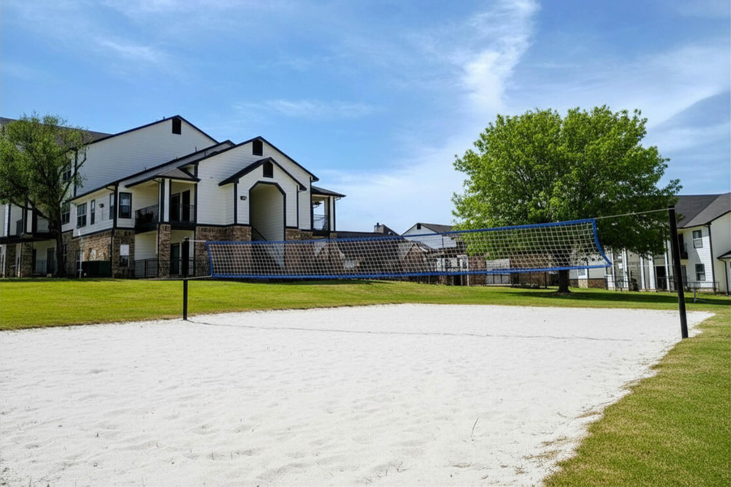 A sandy volleyball court set in a green area with modern apartment buildings in the background. The sky is clear with a few clouds, and a large tree provides shade nearby. The scene is inviting and suitable for outdoor activities.
