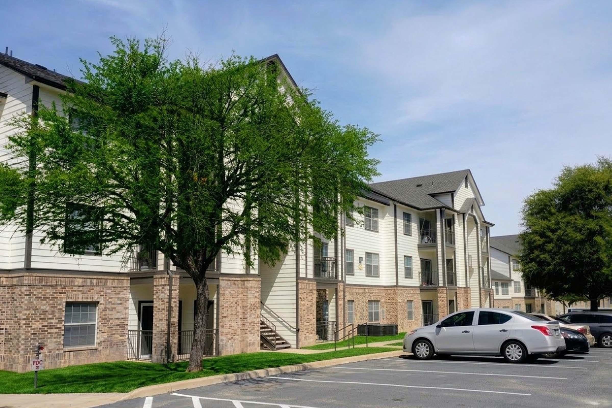 A view of a multi-story apartment building featuring a mix of brick and siding. The building has balconies, grassy areas, and several parked cars in the foreground. Trees provide shade near the entrance, with a clear blue sky above.