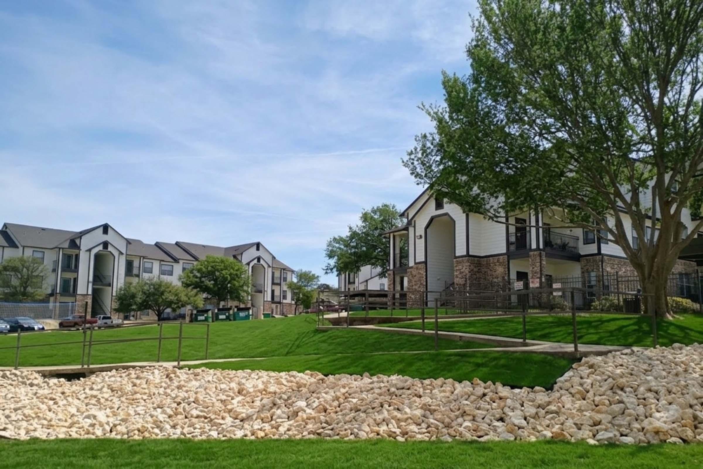 A landscaped area featuring green grass, a rocky border, and modern apartment buildings in the background under a clear blue sky. Trees are visible, adding to the scenic view of the community space.