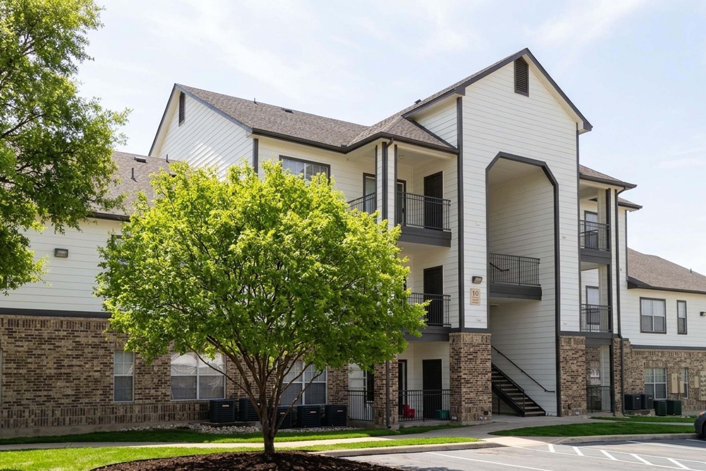 A modern multi-story apartment building featuring a mix of brick and siding exteriors. The building has balconies, a staircase leading to the upper floor, and is surrounded by green landscaping, including a mature tree. The scene is set under a clear blue sky.