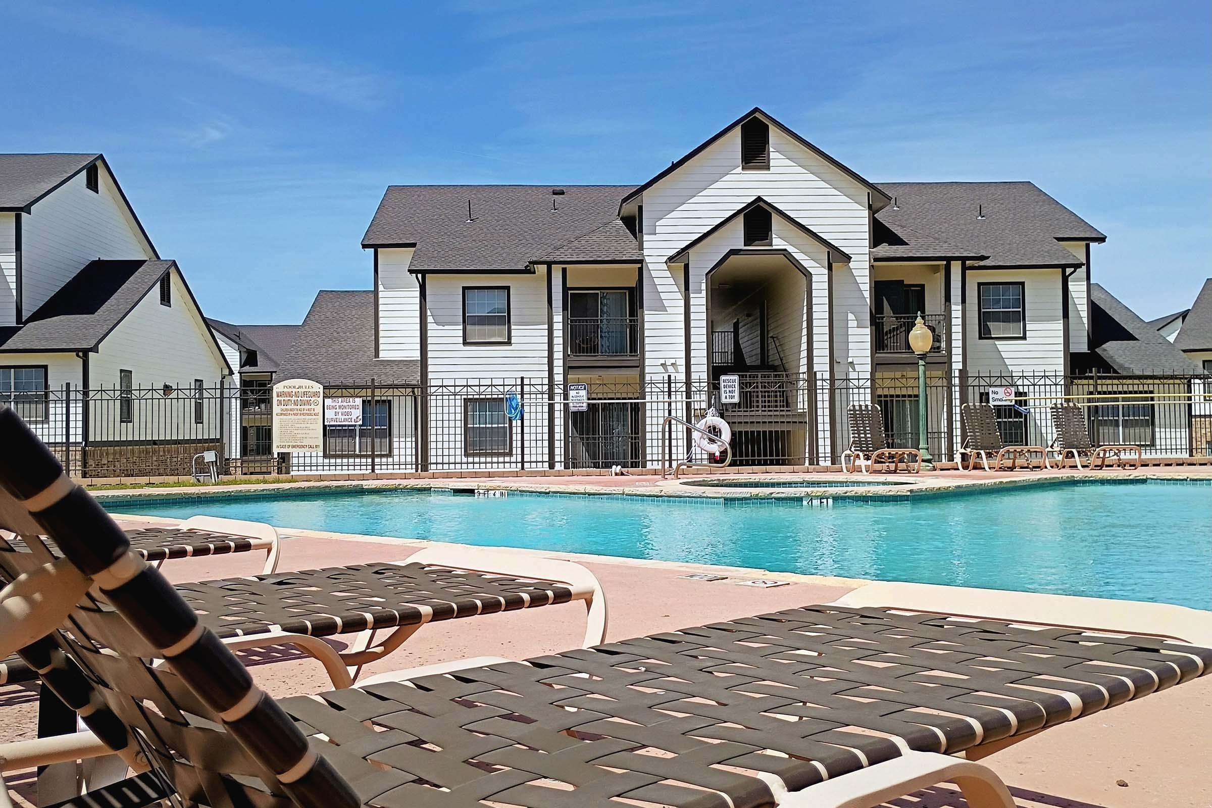 View of a swimming pool surrounded by lounge chairs, with an apartment building in the background. The pool area is fenced and includes signs, and there are clear blue skies overhead. The scene suggests a relaxing residential environment.