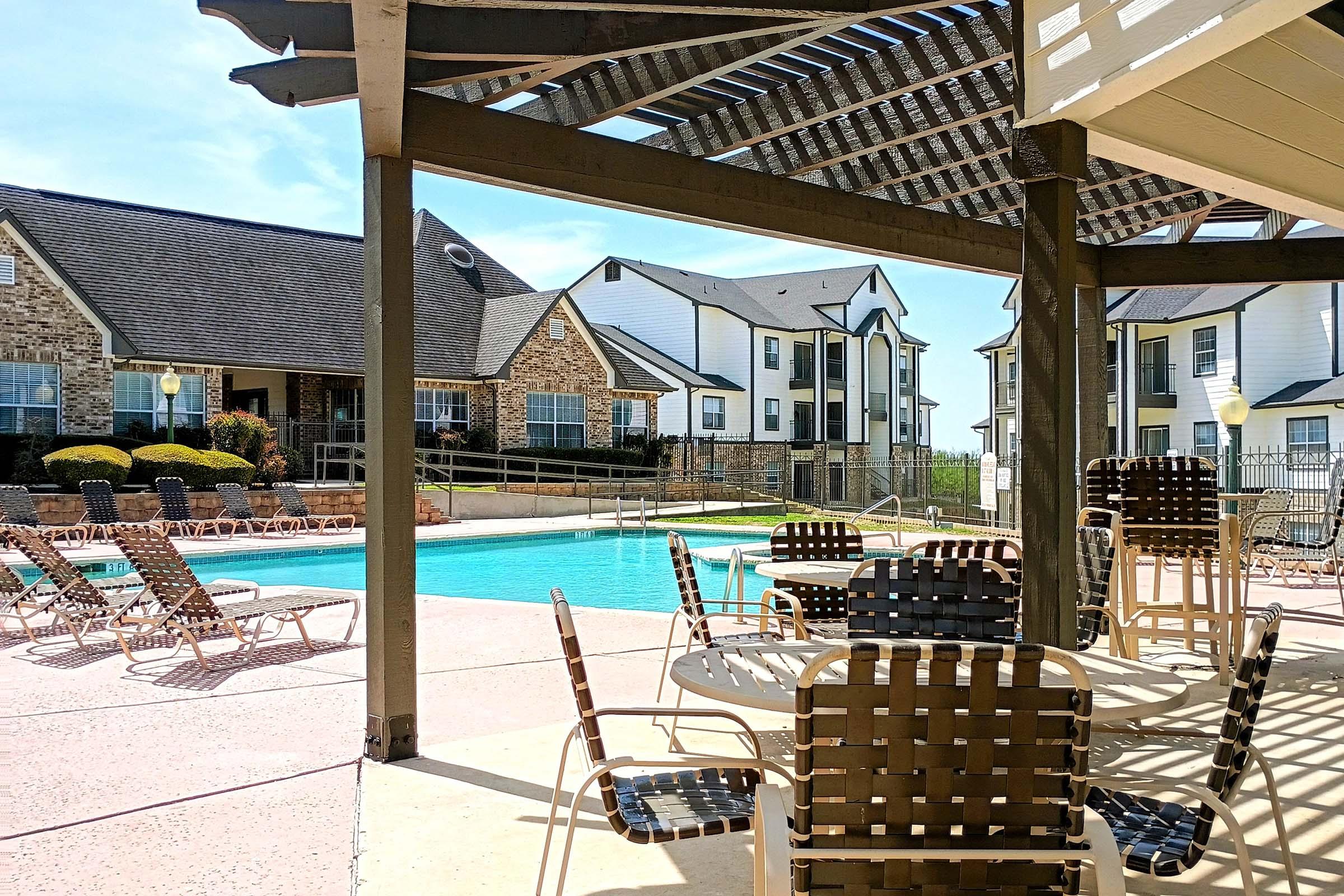 A sunny pool area featuring a large swimming pool surrounded by lounge chairs and tables. The scene includes a covered patio with a wooden pergola, and multi-story residential buildings in the background. Lush landscaping adds to the inviting atmosphere.