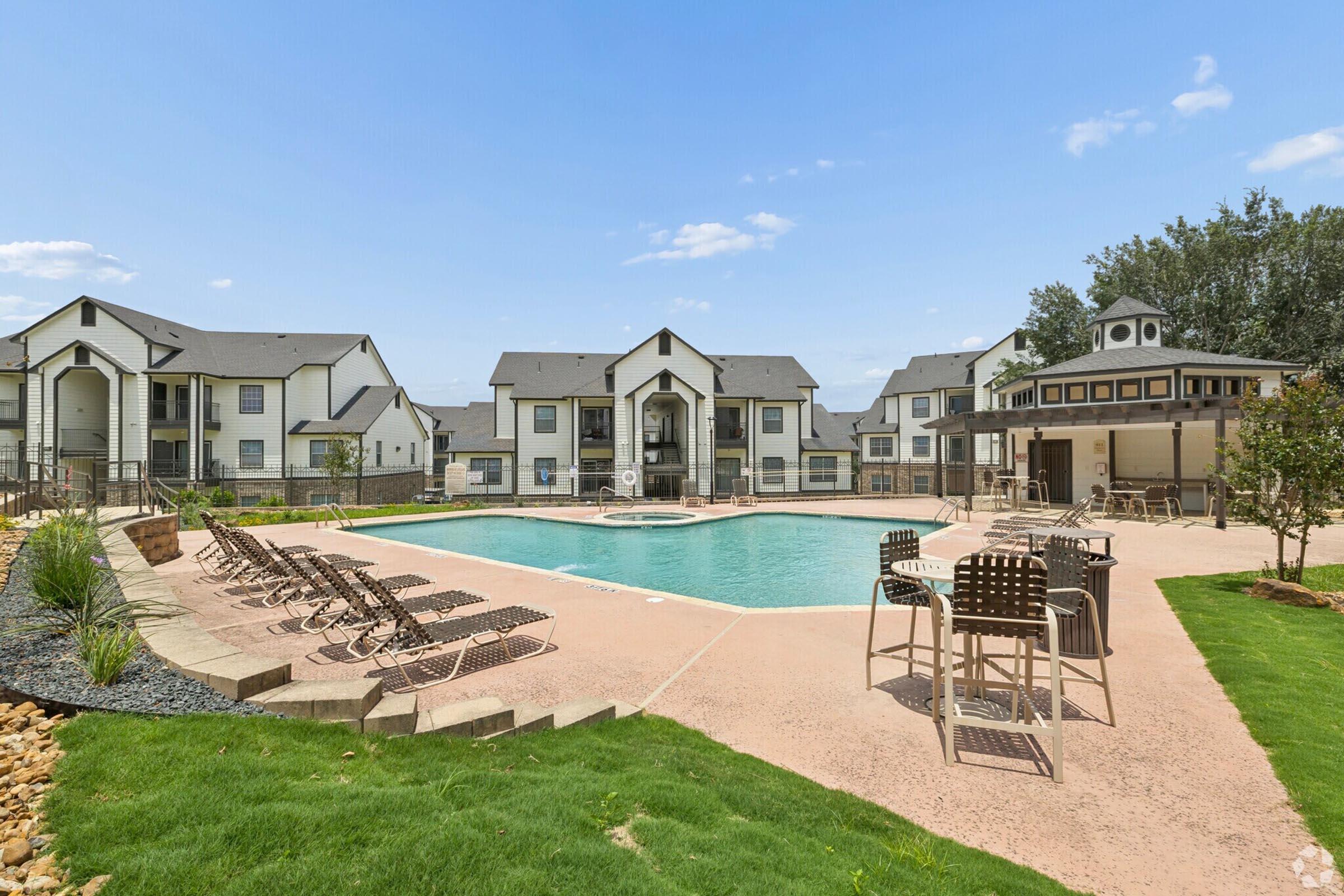 A modern apartment complex featuring a sparkling outdoor swimming pool surrounded by lounge chairs. Lush green grass and landscaped areas are present, with multiple buildings in the background, creating a welcoming community atmosphere. The sky is clear with a few clouds.