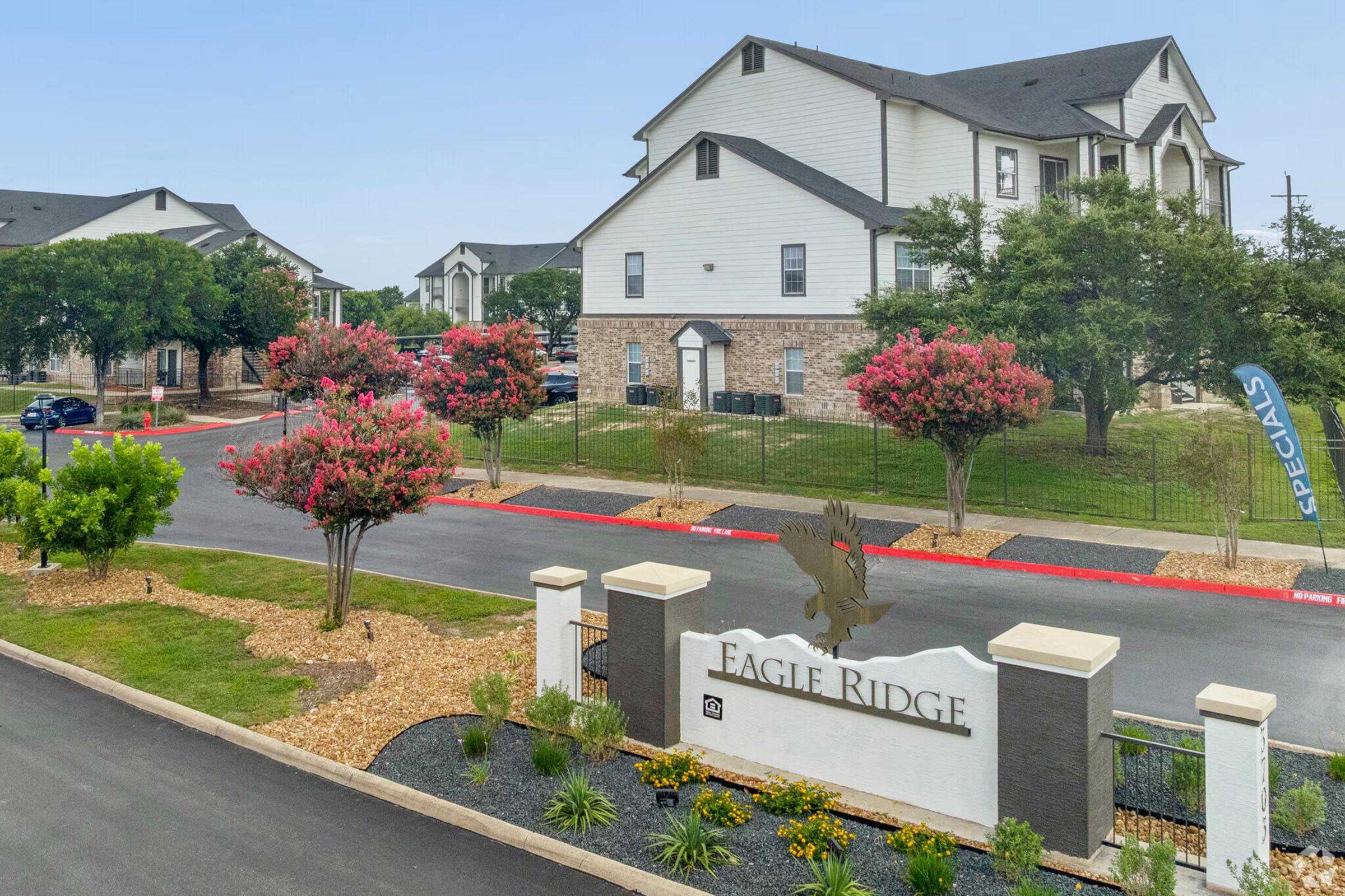 View of the Eagle Ridge apartment complex, featuring a stone and white building with multiple stories. The entrance sign reads "EAGLE RIDGE," surrounded by landscaped flower beds and trees. The road is lined with decorative shrubs, and a promotional banner is visible in the background.