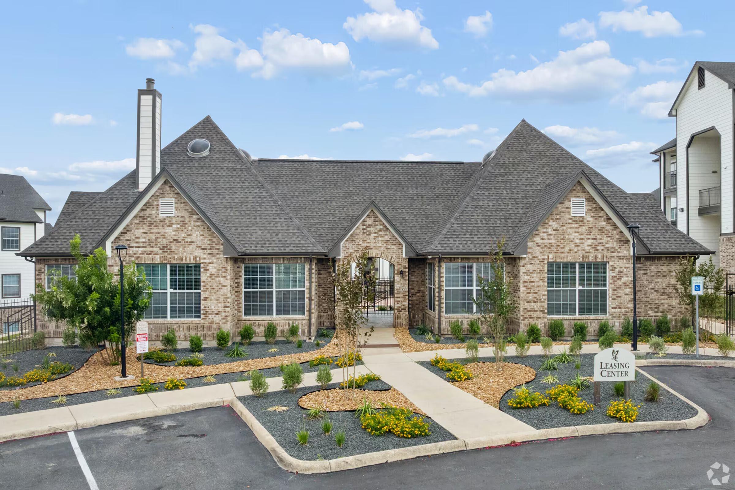 A modern brick leasing center building with a gabled roof, large windows, and landscaped gardens. The entrance features a pathway lined with plants and decorative stones, and there are signs indicating the leasing office. The sky is partly cloudy, adding to the inviting atmosphere.