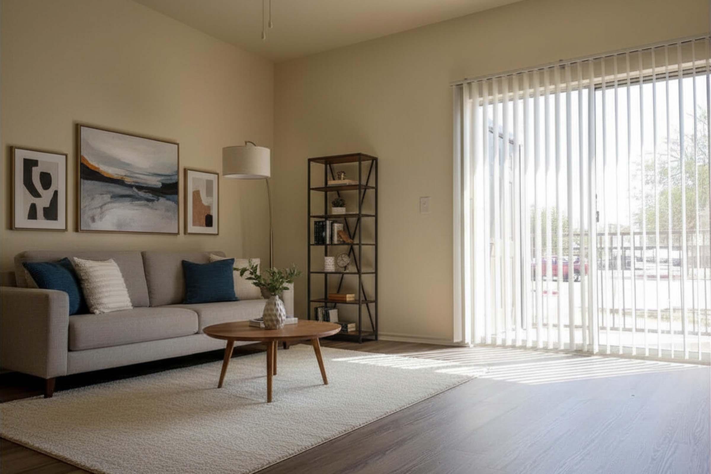 A cozy living room featuring a light gray sofa adorned with blue accent pillows, a round wooden coffee table, and a textured rug. There's a modern bookshelf in the corner and large vertical blinds letting in natural light from a window. Wall art adds a stylish touch to the space.