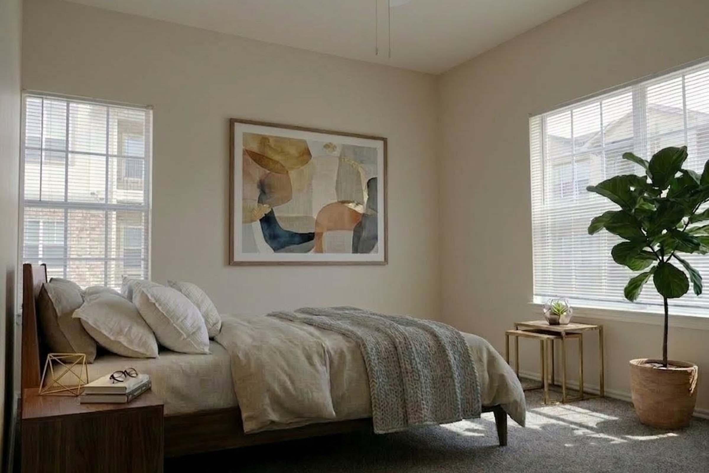 A bright and inviting bedroom with large windows allowing natural light. The room features a bed with fluffy pillows and a soft blanket, a framed abstract art piece on the wall, a small wooden side table with decorative items, and a potted plant adding a touch of greenery to the space.