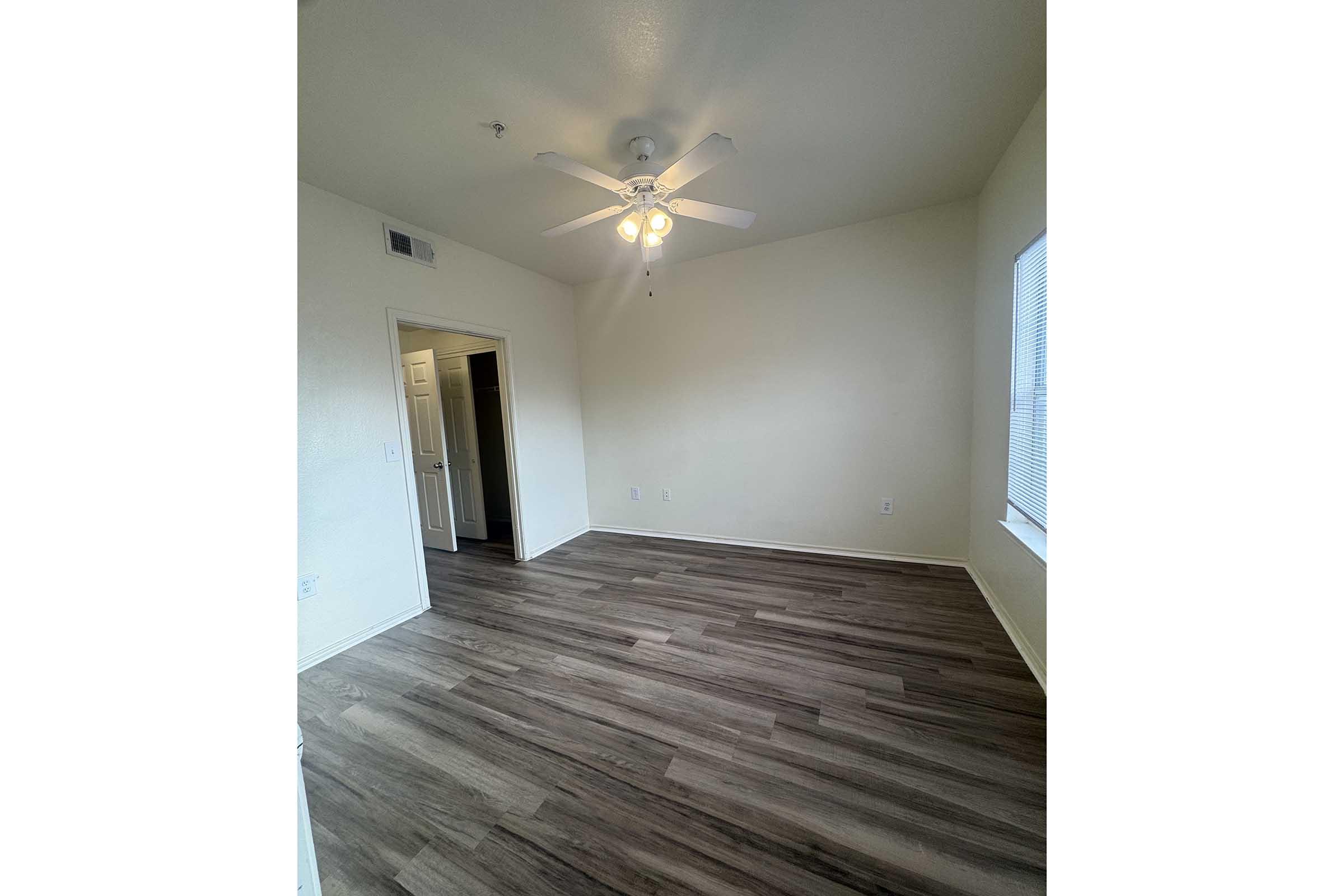 A sparsely furnished room featuring light-colored walls, a ceiling fan with light fixtures, and a window. The flooring is a light wood or laminate style, and there is a doorway leading to another room. The space appears bright and inviting, ideal for various uses such as a bedroom or office.