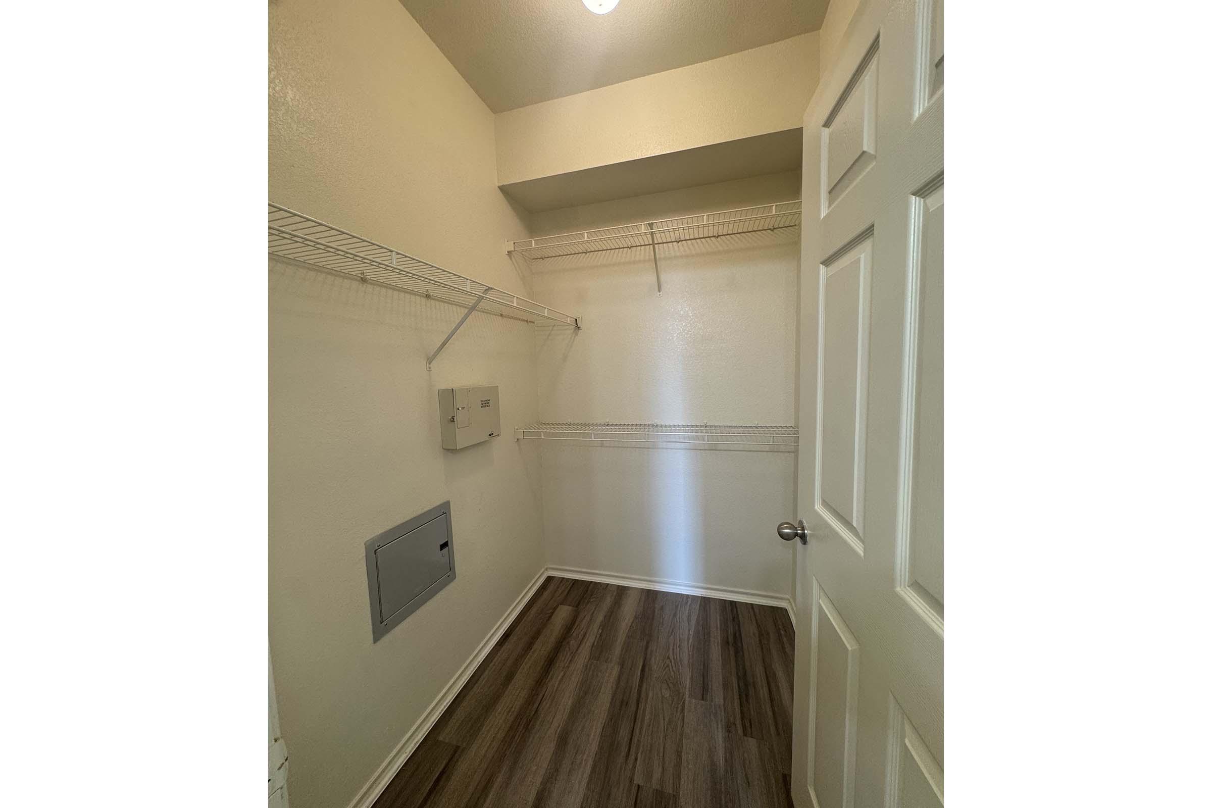 Empty closet with light-colored walls and wooden flooring. The space features wire shelving on the left and back walls, and a small panel on the right side. A door leads to the hallway, and overhead lighting is present.