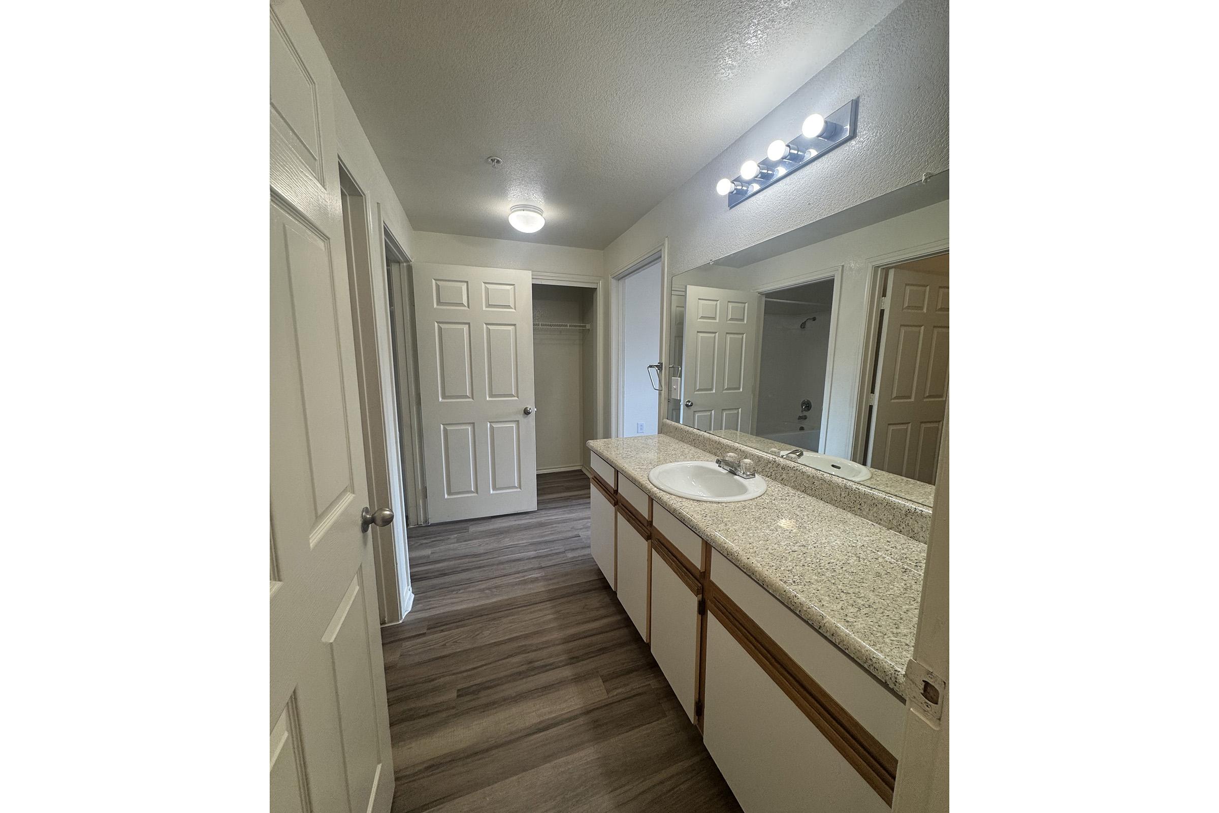 A well-lit bathroom featuring a double vanity with two sinks, granite countertop, and large mirrors above. The room has beige walls, wooden flooring, and a door leading to a closet. The overall design is clean and modern, emphasizing simplicity and functionality.
