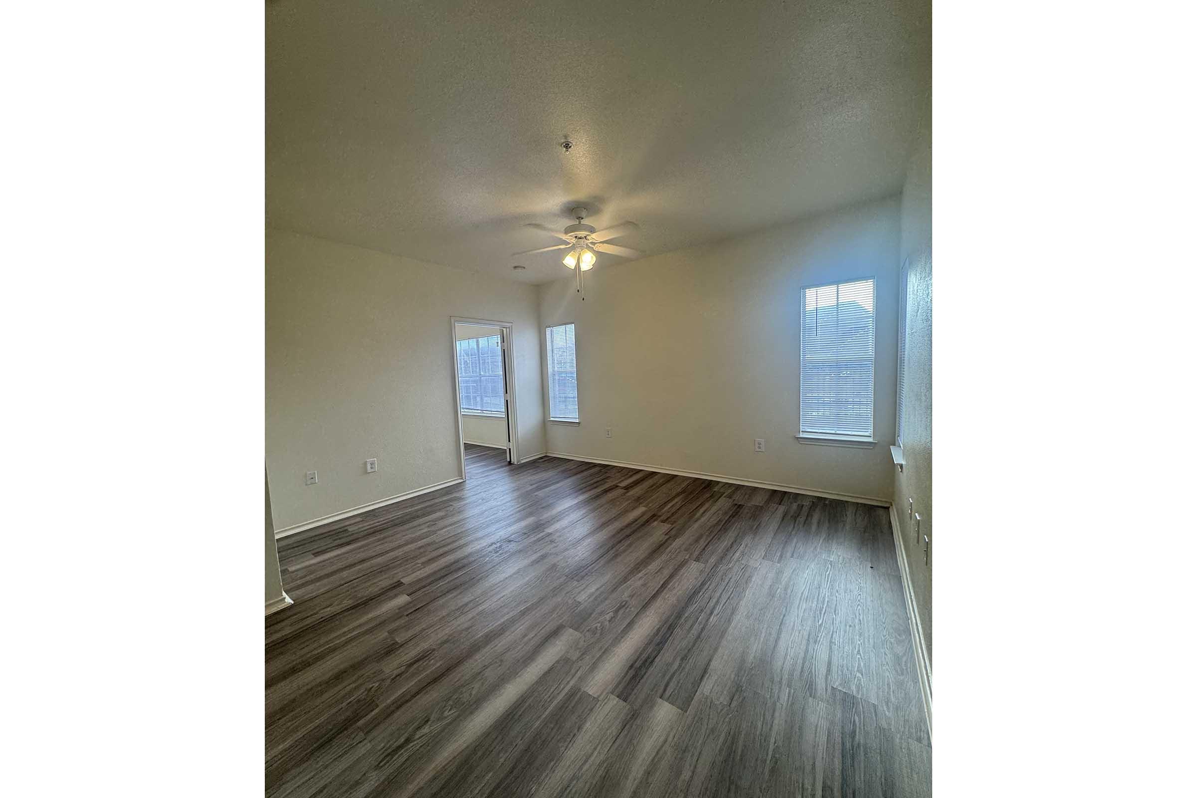 Empty room with light-colored walls and wooden flooring. A ceiling fan is mounted in the center, and large windows provide natural light. One wall features a doorway leading to another space, and the overall ambiance is bright and spacious.