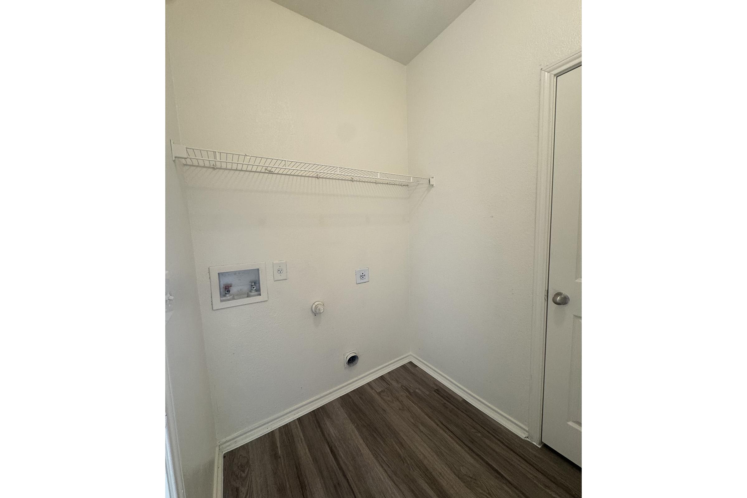 A minimalist laundry room with white walls, featuring a wire shelf mounted on one side. Electrical outlets and connections are visible on the wall, while the floor has a wood-like laminate finish. A closed door is seen on the right, indicating access to another room.
