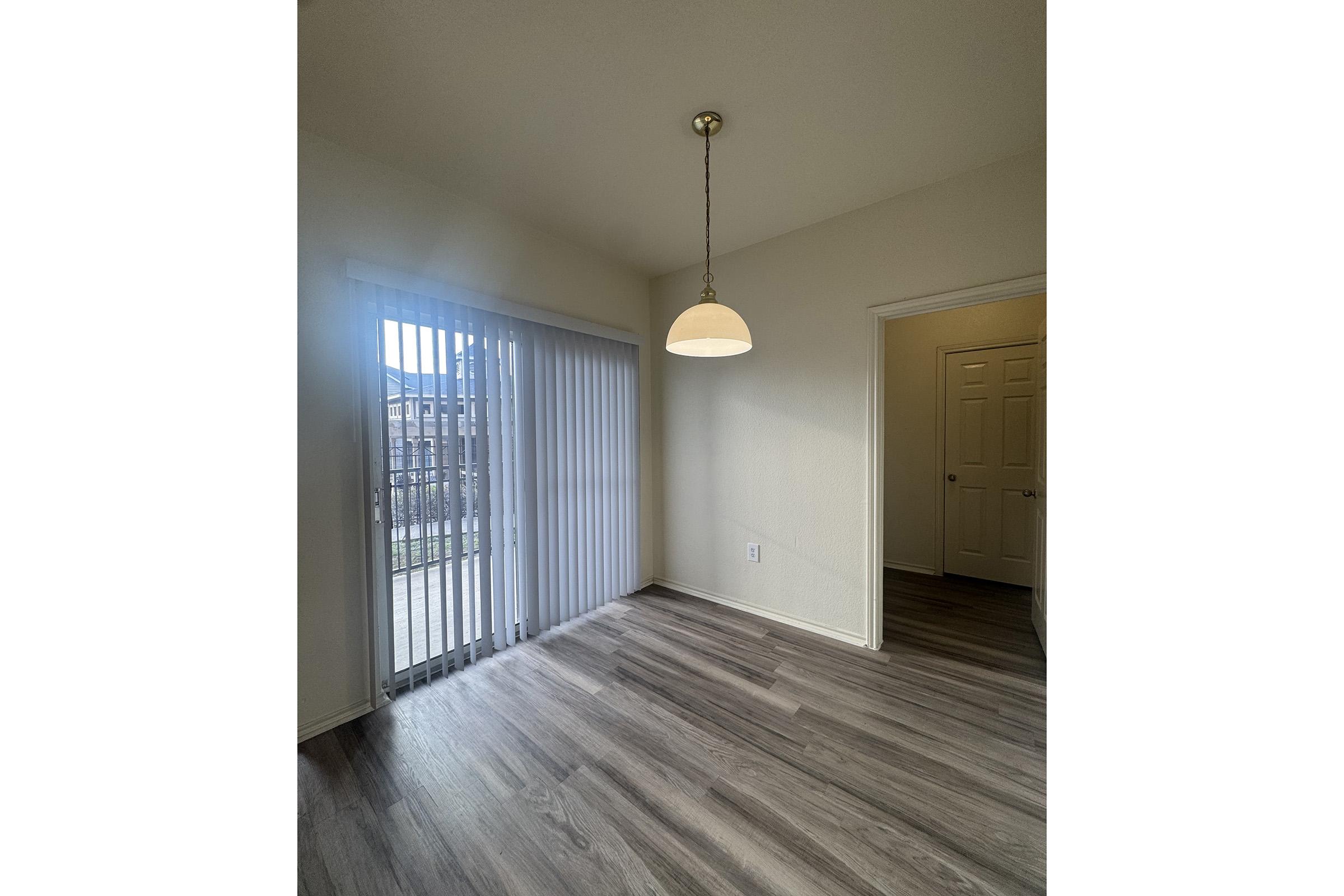 Inside view of a room with wooden flooring, sliding glass doors leading to an outdoor area, and a ceiling light fixture. The walls are painted a light color, creating a bright and open atmosphere. A doorway on the right leads to another room.