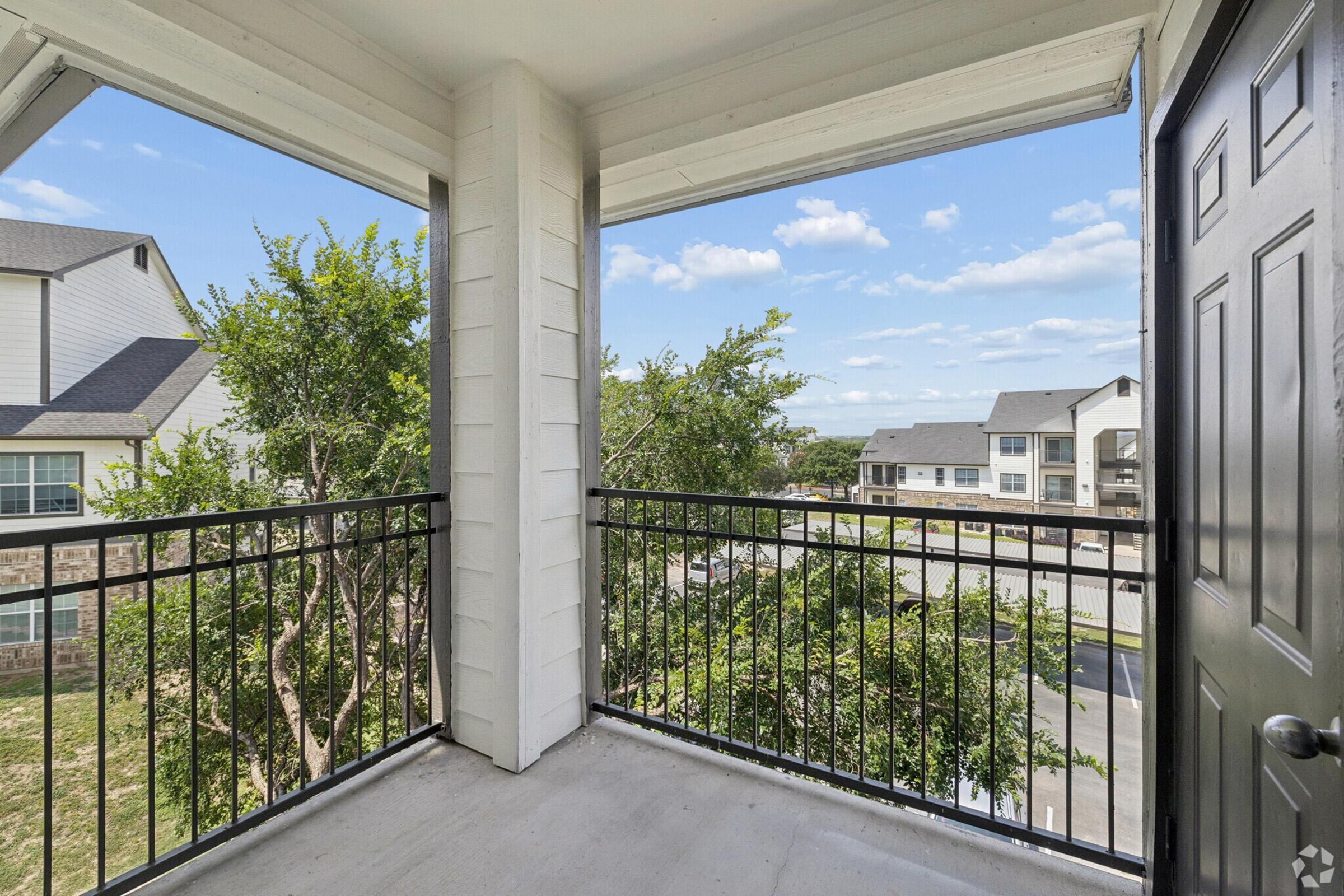 View from a balcony featuring a black railing, overlooking green trees and buildings in the distance. The sky is clear with a few clouds, and the structure has a modern design with light-colored walls. The scene conveys a sense of openness and natural light.