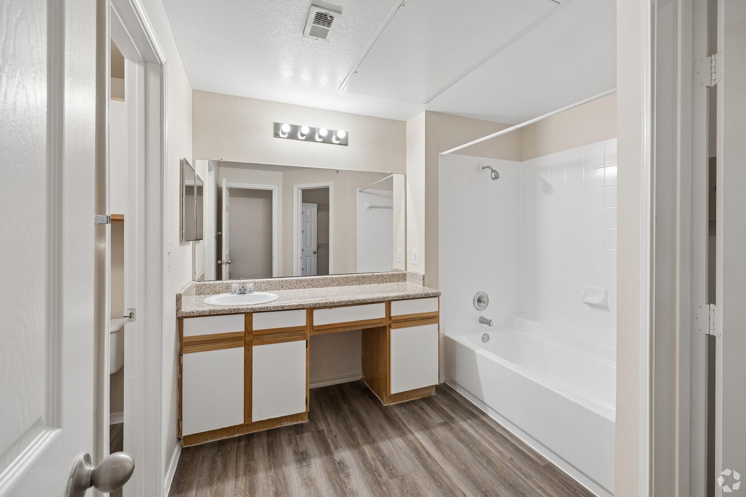 A bathroom featuring a white tub and shower combination, a double sink vanity with a granite countertop, a large mirror above the sinks, and light fixtures. The walls are painted in a neutral tone, and the floor has brown laminate flooring. There is a door leading to another room in the background.