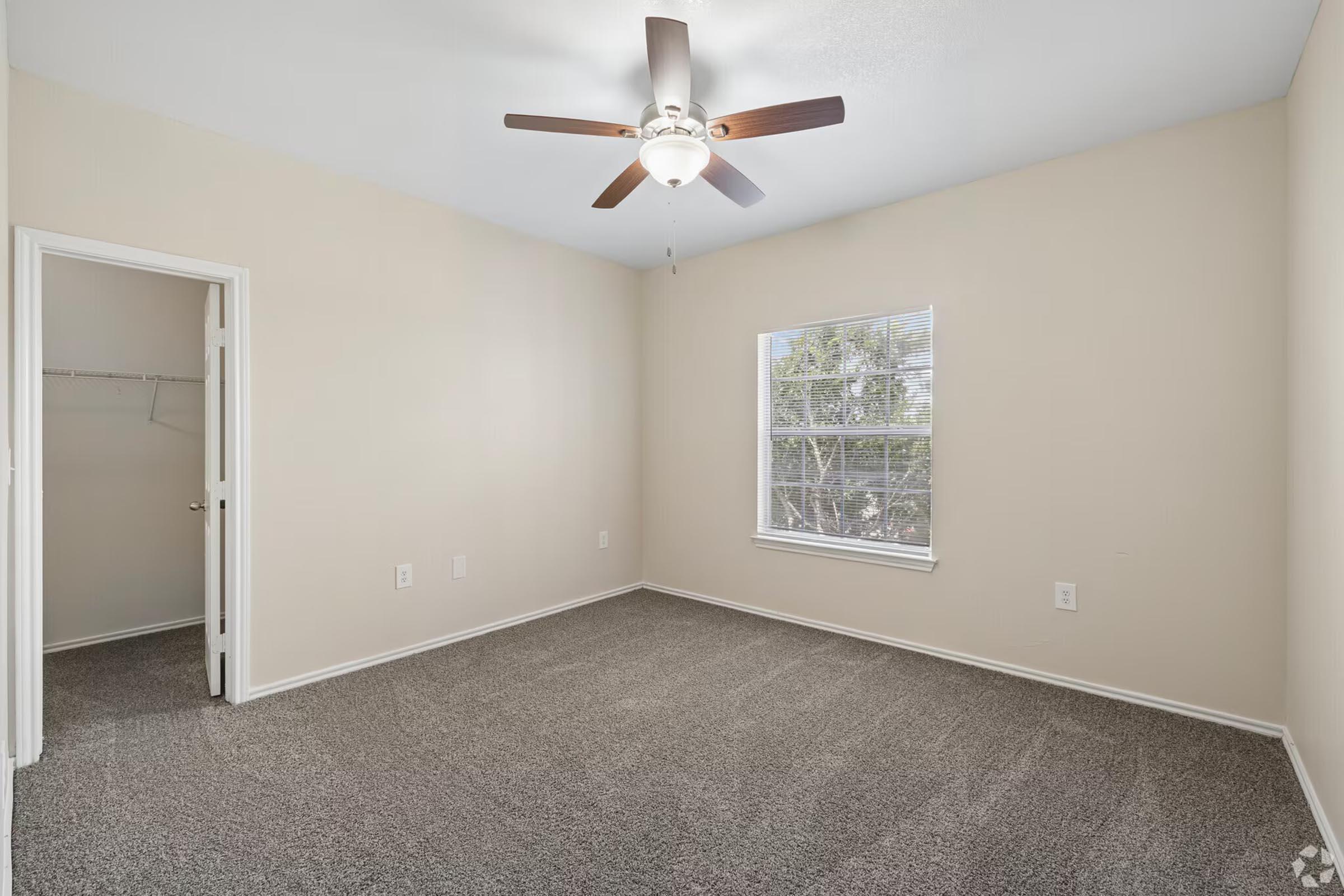 A vacant bedroom with light beige walls and gray carpet. It features a ceiling fan with a light fixture and a window allowing natural light. A closet is visible in an adjacent room through an open door, creating a spacious and airy feel.
