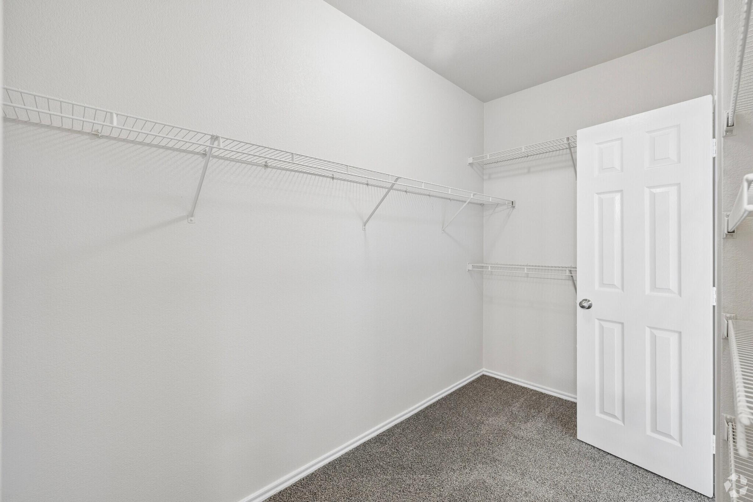 An empty closet featuring light-colored walls, a beige carpeted floor, and wire shelving along two walls. A white door is closed, and the space appears clean and organized, suggesting potential for storage or personal organization.