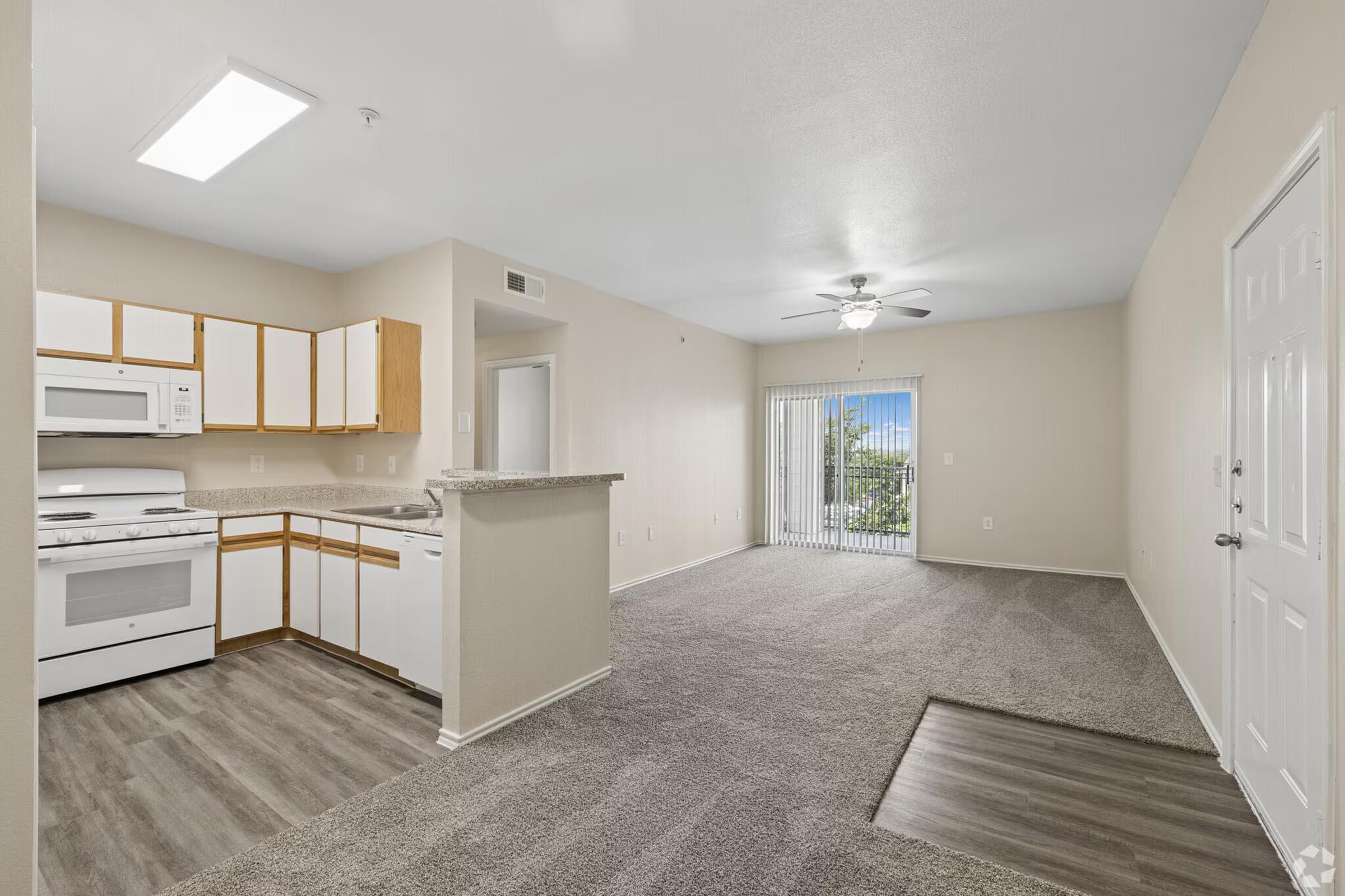 A spacious apartment interior featuring a kitchen with white cabinets and appliances, a living area with carpet flooring, and a sliding glass door leading to a balcony. Natural light enters through the door, creating a bright and inviting atmosphere. Walls are painted in neutral tones.
