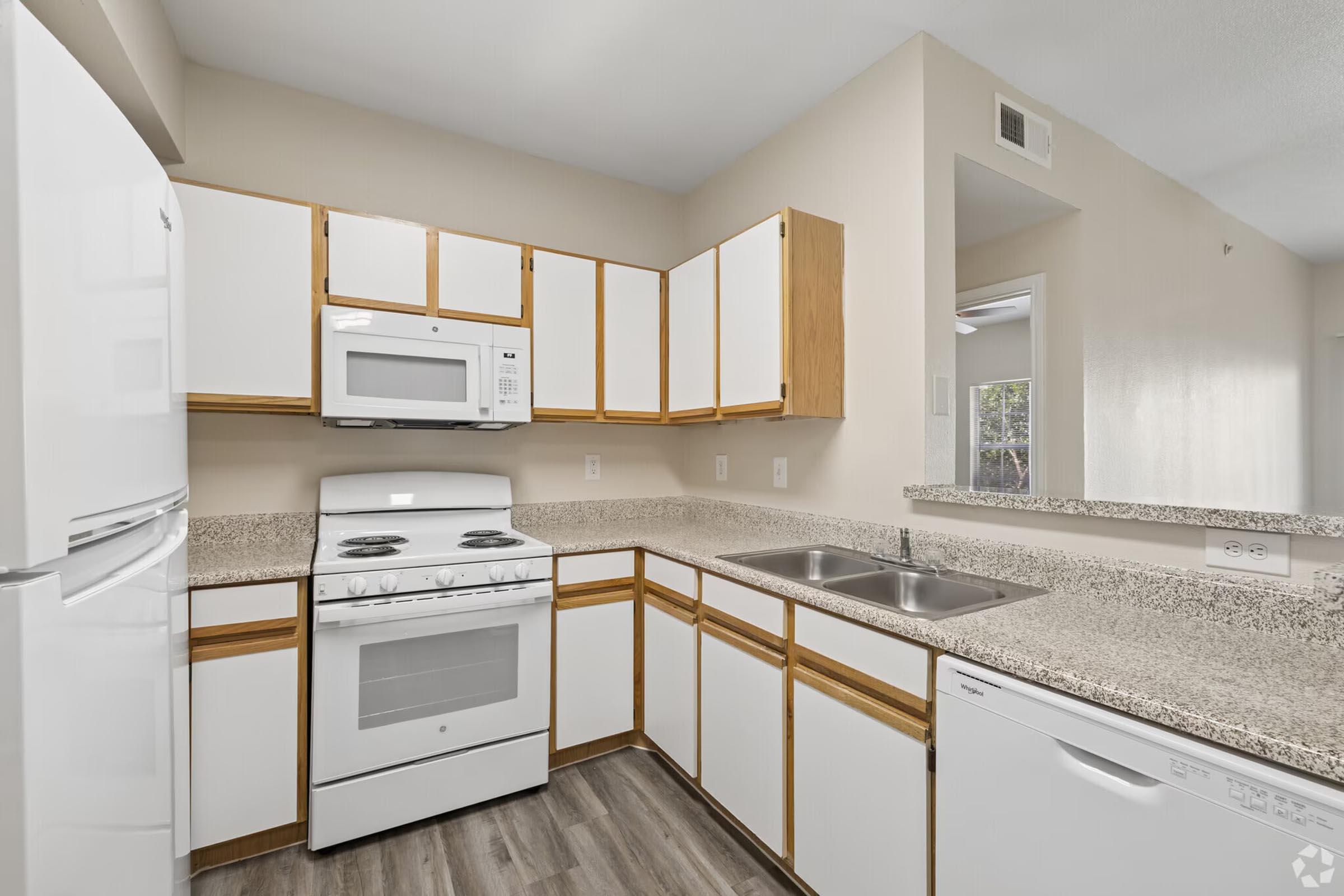 A clean and modern kitchen featuring white cabinets with wooden accents, a white refrigerator, a stove with an oven, a microwave, and a double sink. The countertops are light-colored and speckled, with a window visible in the background, and a seamless flow into the adjacent area.