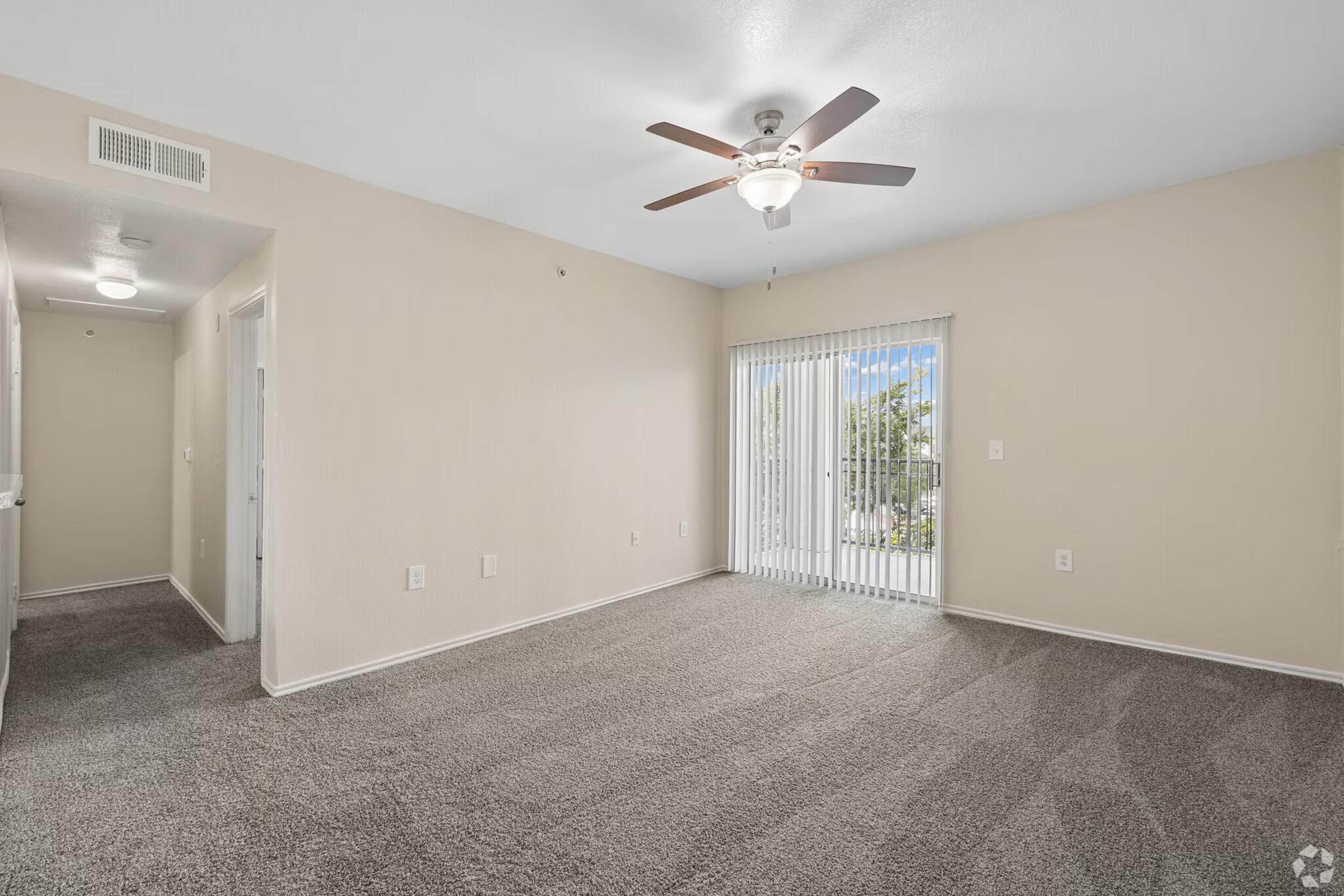 Empty living room with light beige walls and plush gray carpet. A ceiling fan is installed above, and sliding glass doors lead to the outside, allowing natural light to enter. The room has a spacious feel with no furniture, creating an inviting atmosphere.