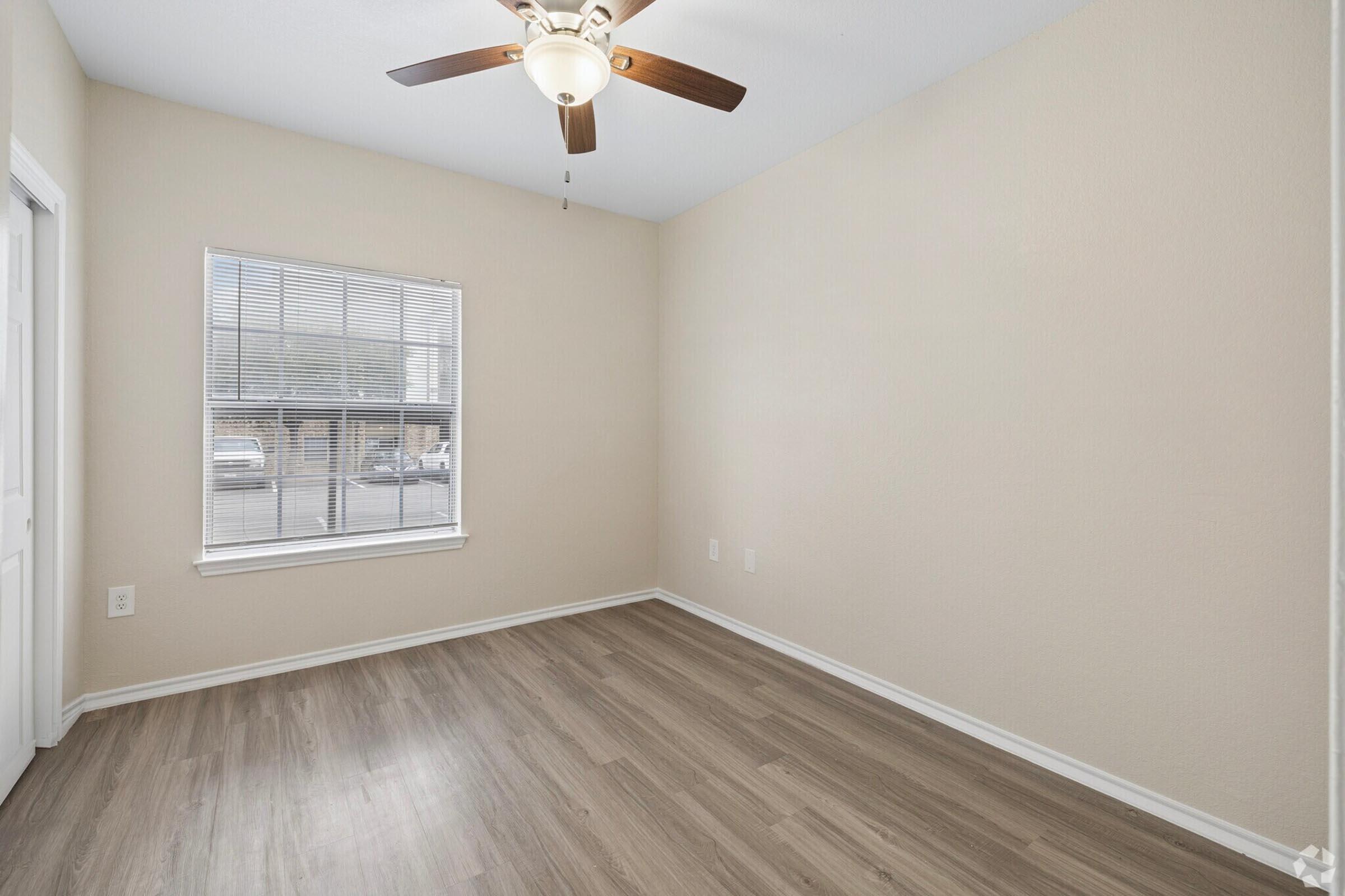 A small, empty room with light beige walls and wooden flooring. A ceiling fan with wooden blades is visible. There’s a window letting in natural light, and the room has minimal furniture, creating a spacious feel. The door is on the left side of the image.