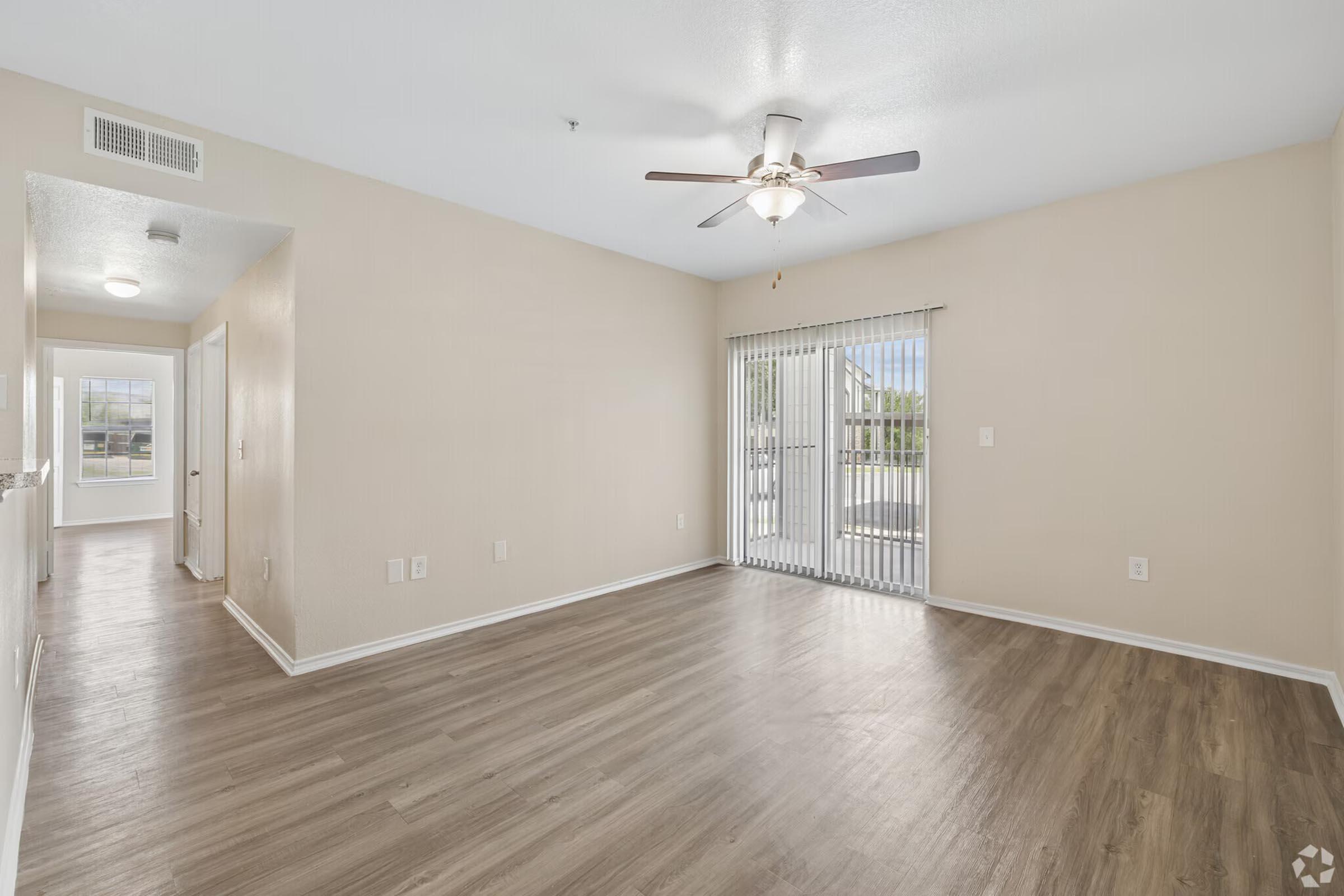 A spacious living area featuring light-colored walls, a ceiling fan, and laminate flooring. There is a sliding glass door with vertical blinds leading to an outdoor area. The room is well-lit with natural light coming in, creating an inviting atmosphere. A doorway leads to a hallway on the left.