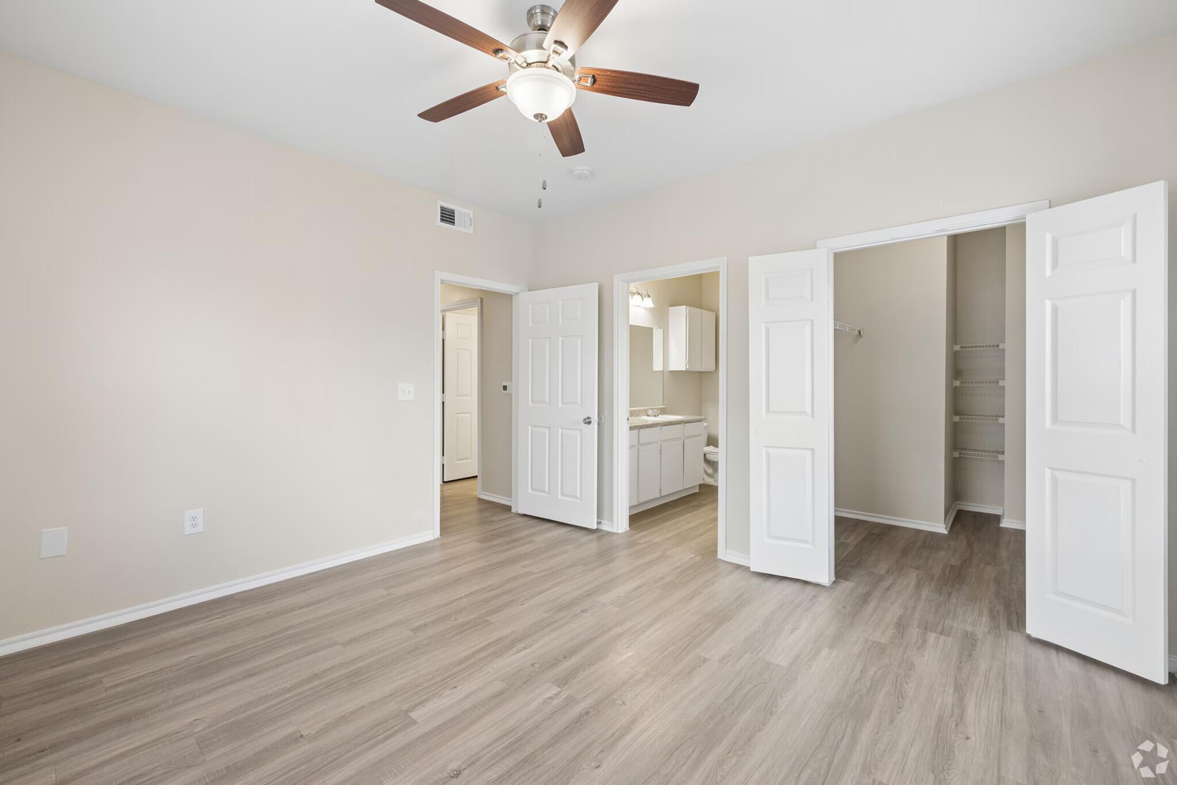 A spacious room featuring light-colored walls and hardwood-style flooring. It includes a ceiling fan and adjoining bathroom, with two open closet doors. The layout offers a bright and airy feel, suitable for various furniture arrangements.