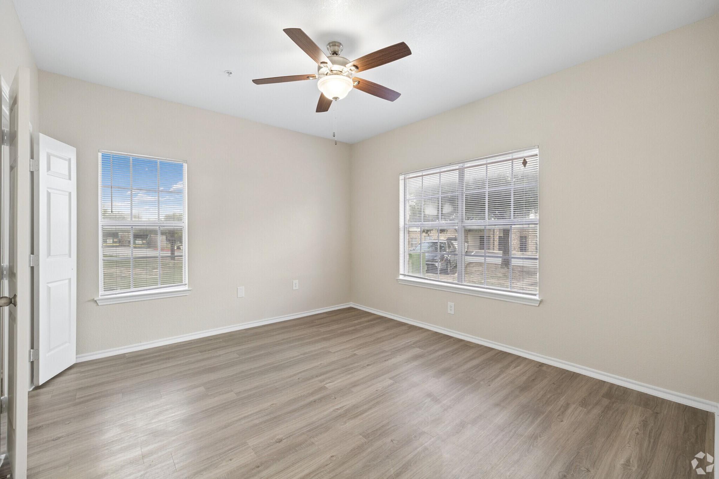 Spacious interior of a room with light-colored walls and wooden flooring. Features a ceiling fan and two windows allowing natural light, with a view of greenery outside. One door is partially open, revealing a simple entrance to the space.