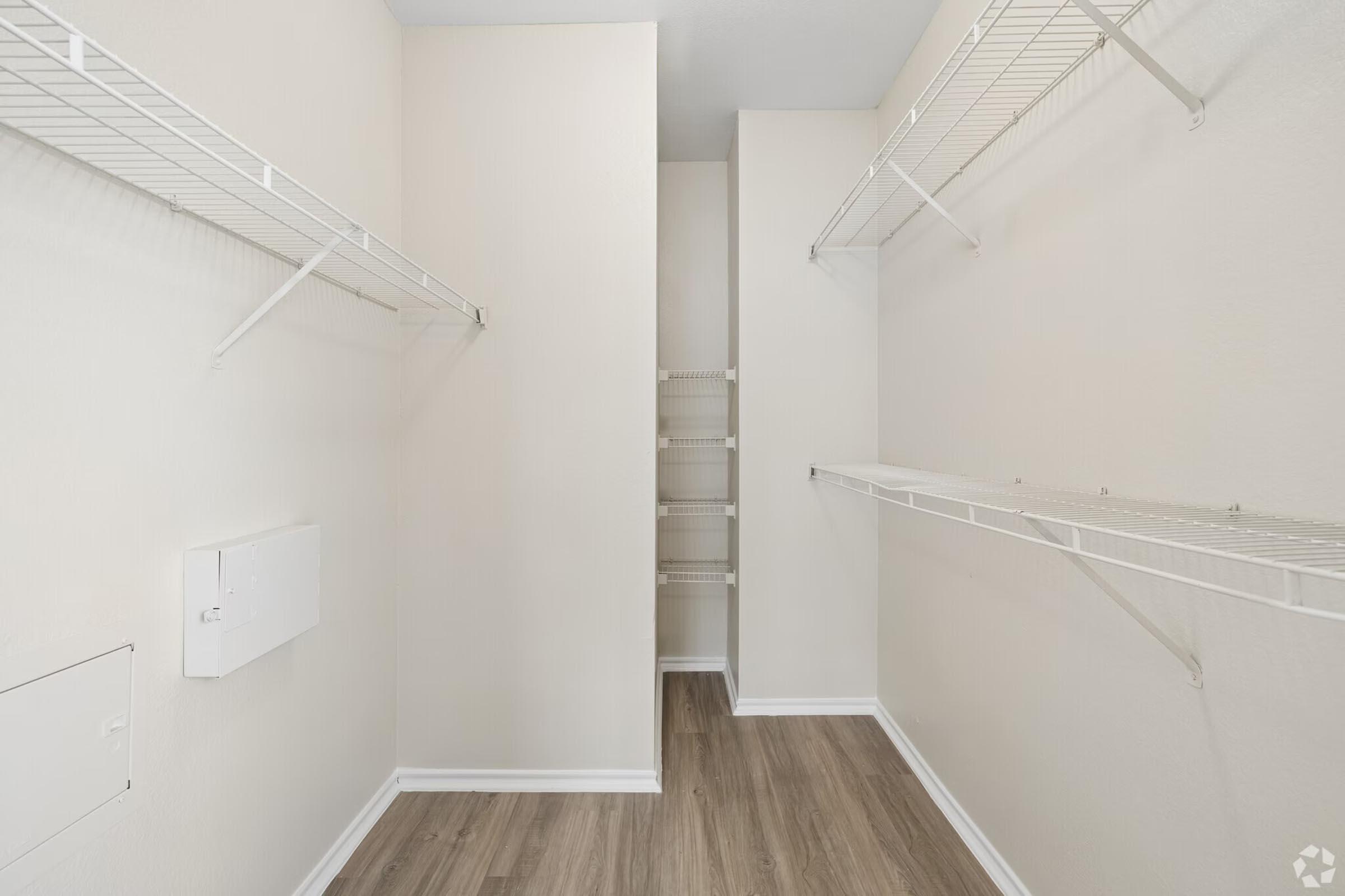 A spacious, empty closet with wire shelving on both sides and a small nook at the back. The walls are painted light color, and the floor has a wood-like finish. There is a white electrical panel on the left side of the wall.