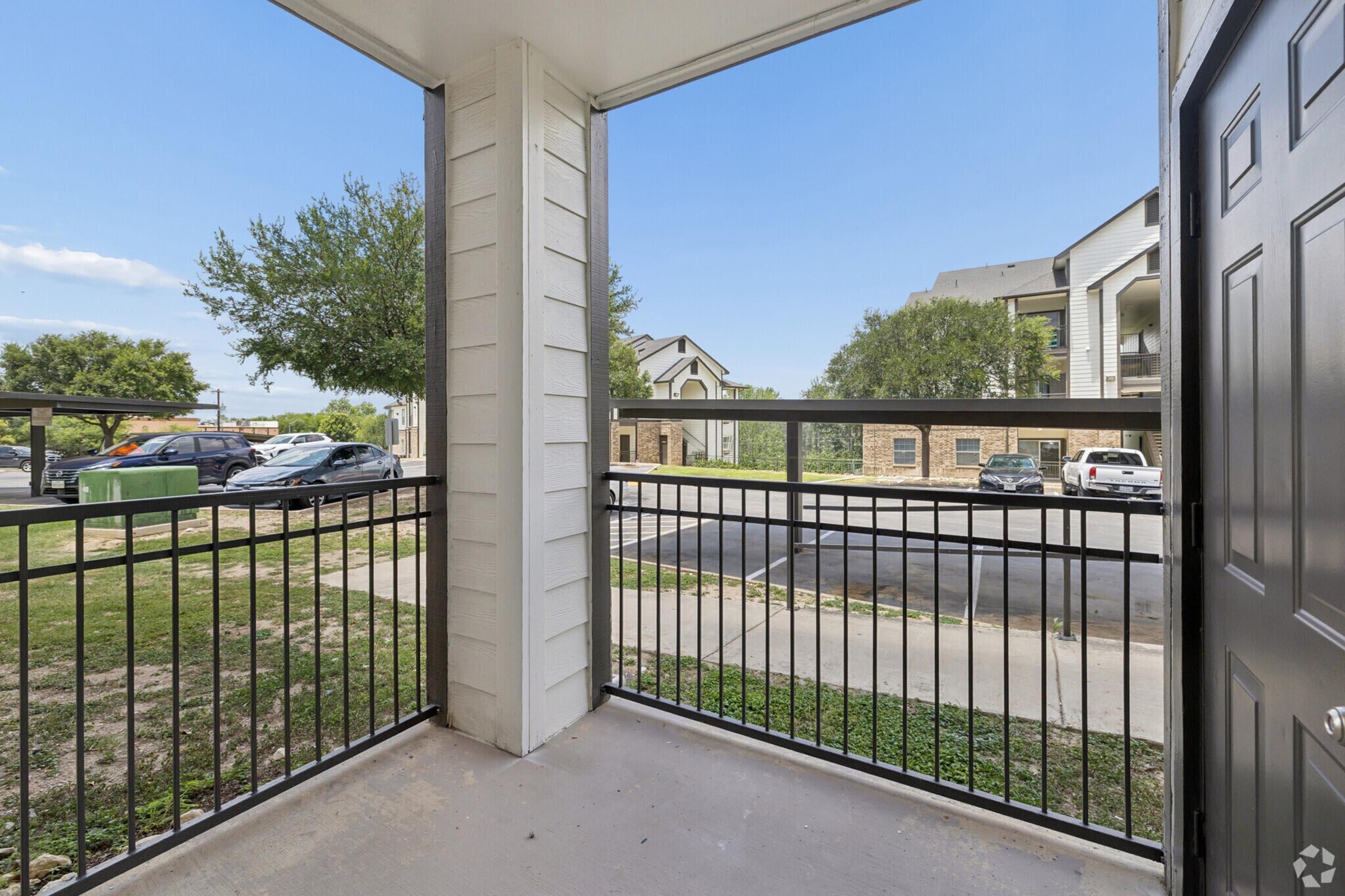 View from a balcony overlooking a green grassy area, with a parking lot and cars visible in the background. The balcony features black railing and is part of a multi-story residential building. The sky is clear and blue, creating a bright and open atmosphere.