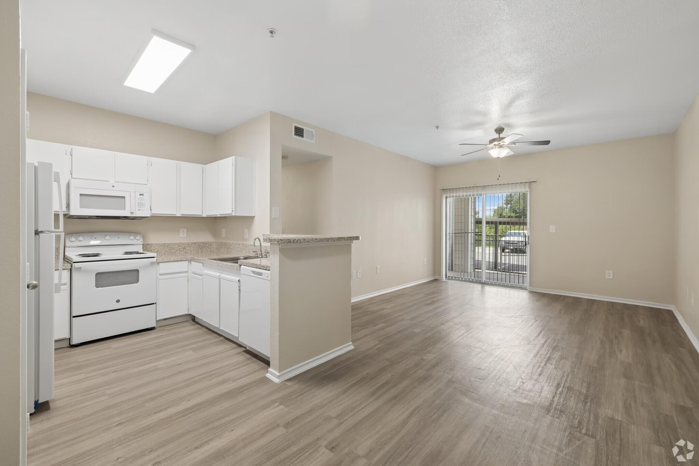 A spacious kitchen and living area featuring white cabinetry, stainless steel appliances, and a ceiling fan. The kitchen includes a breakfast bar and overlooks a bright living room with sliding glass doors leading to a patio or balcony. The flooring is light wood laminate, enhancing the open feel of the space.