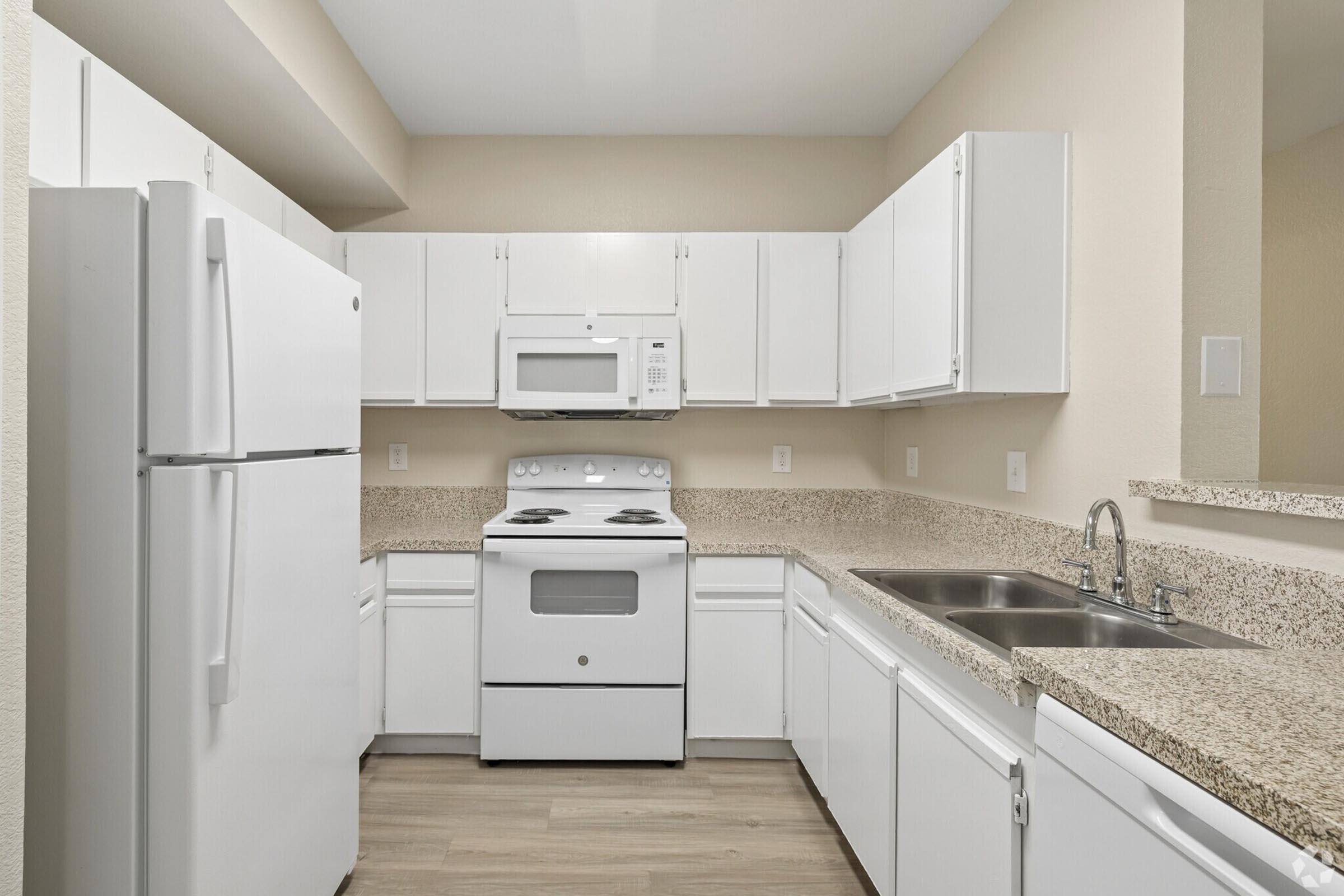 A modern kitchen featuring white cabinets, a white refrigerator, a white microwave above the stove, and a double sink. The countertops are a light-colored granite. The kitchen is well-lit and has a neutral wall color, creating a clean and spacious appearance.
