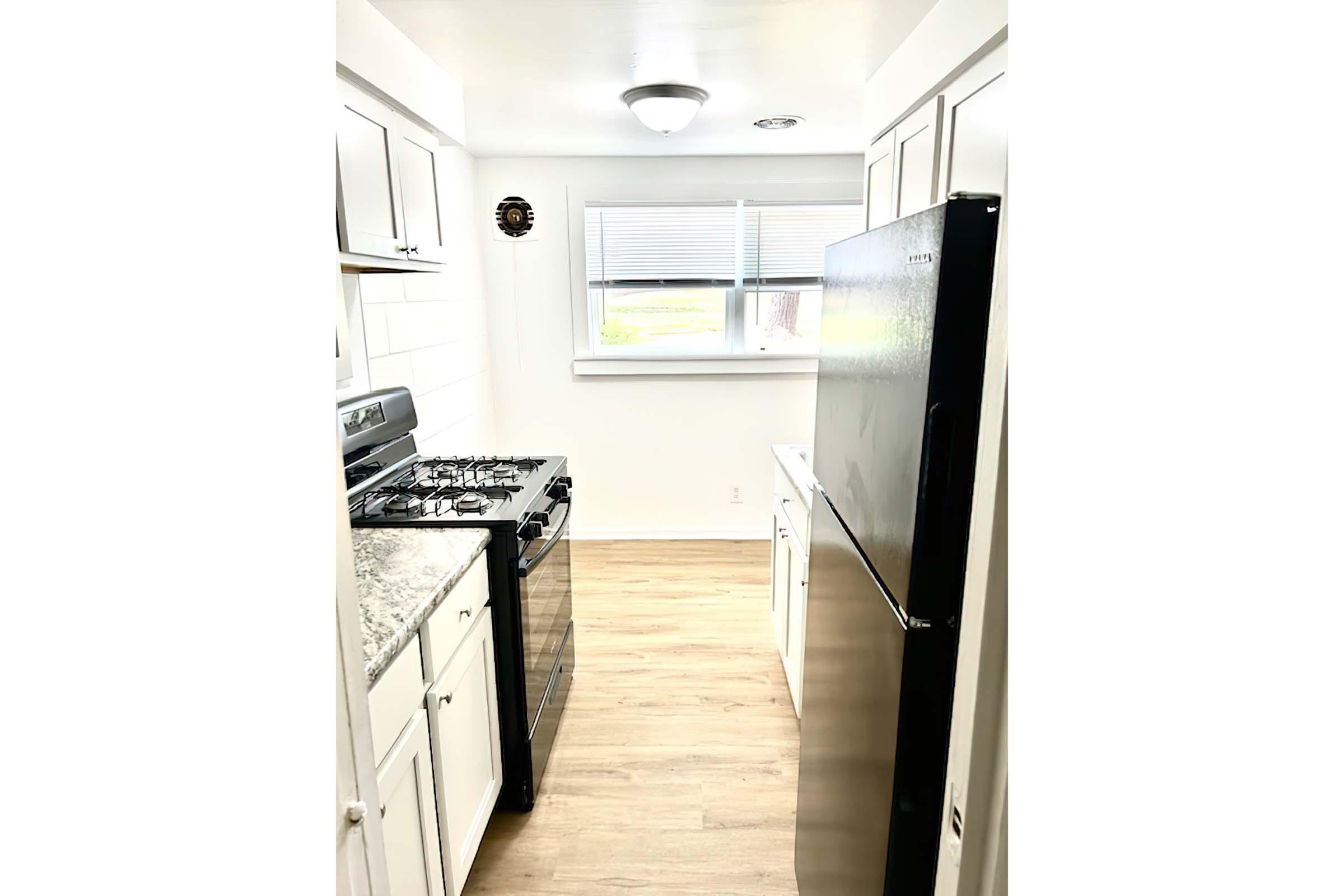 A modern kitchen featuring a black refrigerator, a gas stove with an oven, and white cabinetry. The walls are finished in light colors, and there is a window providing natural light. The flooring is light wood, enhancing the contemporary look of the space.