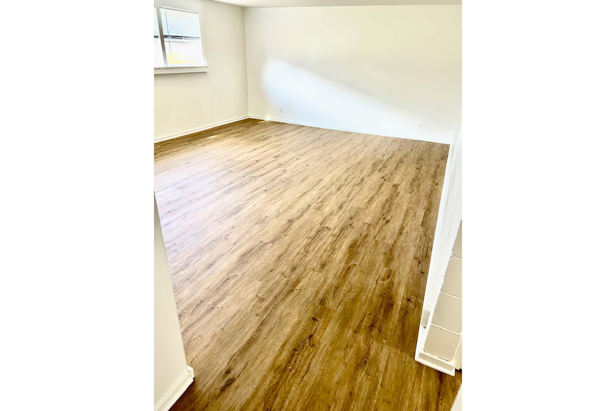 A bright, empty room with light-colored walls and a large window. The floor is covered with light brown laminate or hardwood, giving the space a warm, inviting feel. The room has no furniture, making it appear spacious and ready for decoration.