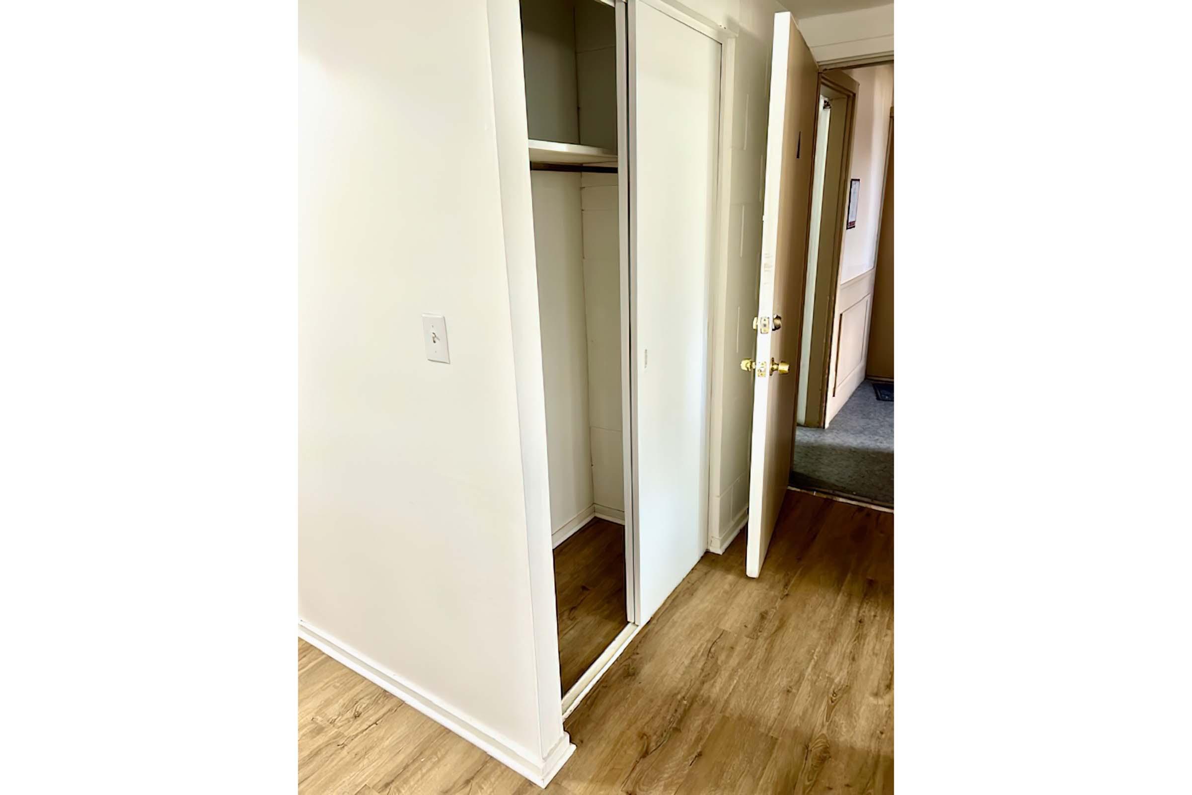 A view of a small, empty closet with mirrored sliding doors, situated beside an open door that leads to a hallway. The walls are painted white, and the flooring is a light wood. The atmosphere is bright and airy, with soft lighting illuminating the space.