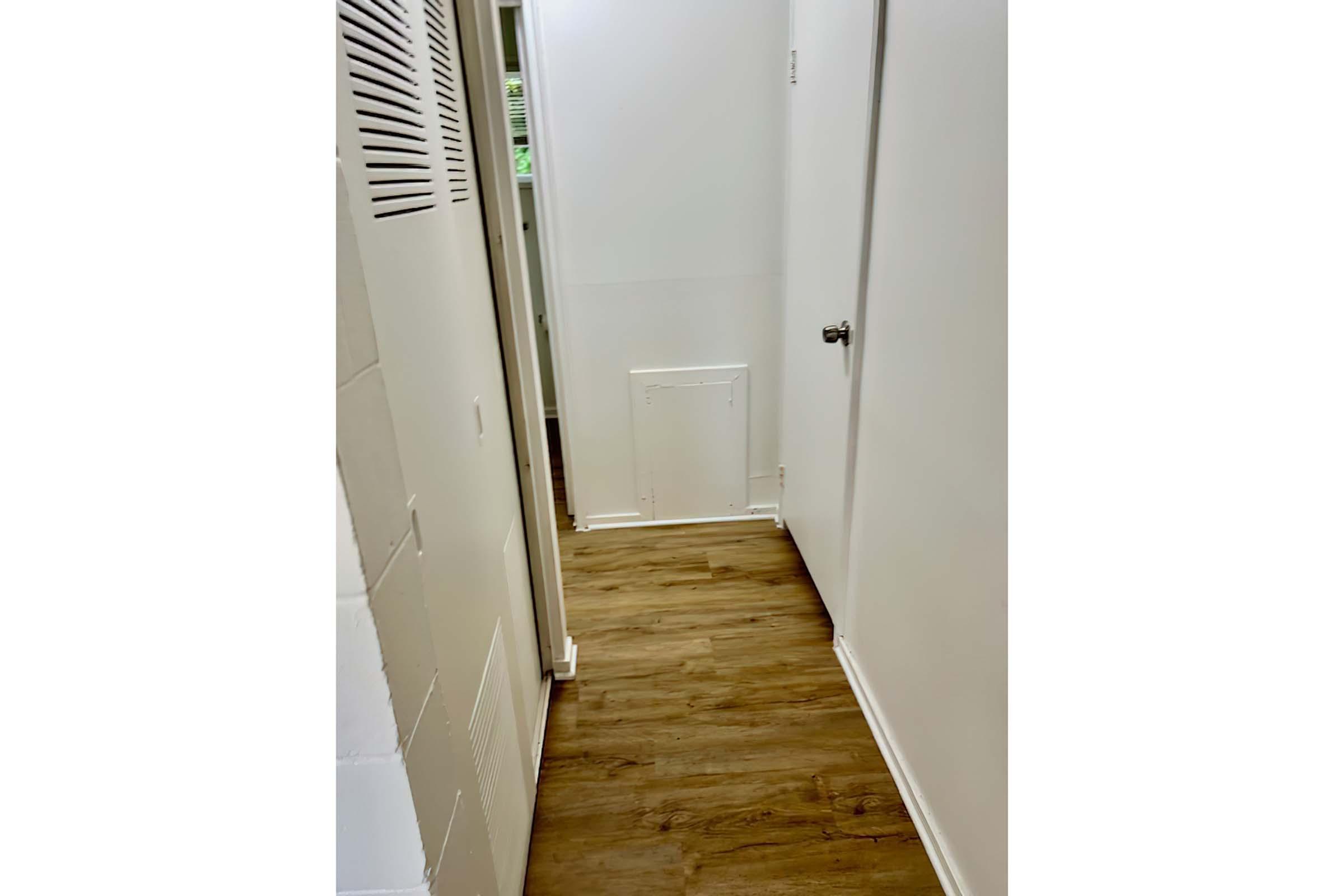 A narrow hallway with white walls and wooden flooring. There is a door on the right and an access panel on the wall towards the end of the hallway. Natural light is visible from a window at the far end, creating a bright atmosphere in the space.